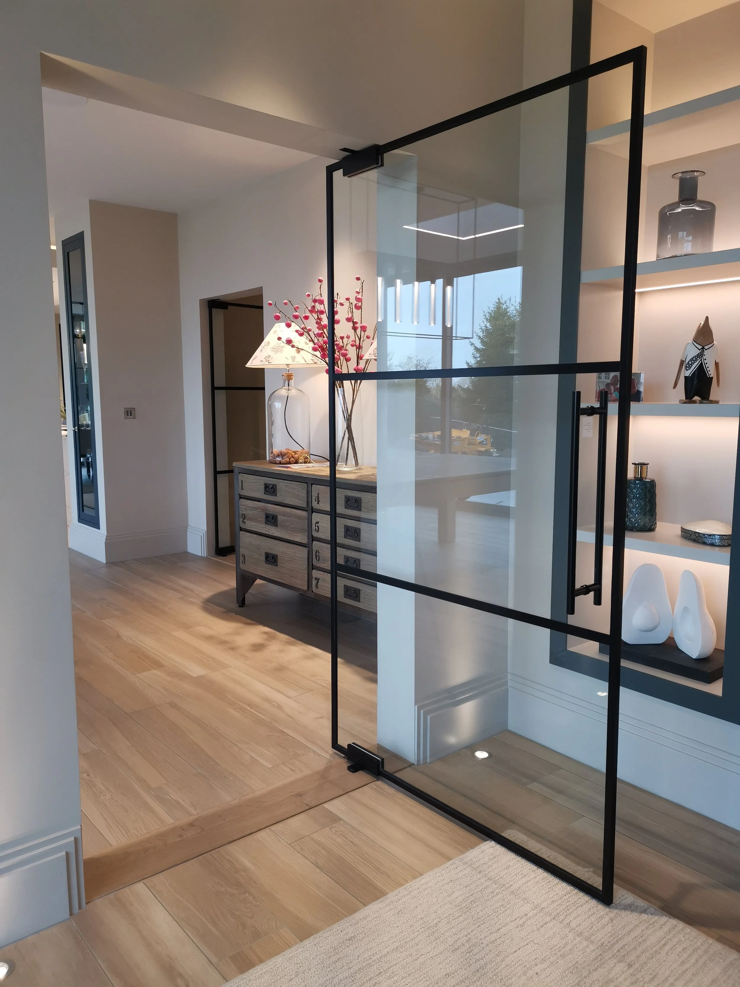 Interior view of a modern home showing a wooden floor, a glass door, a textured white staircase, a dresser with a glass vase and pink flowers, and a decorative lamp and sculptures on a shelf.