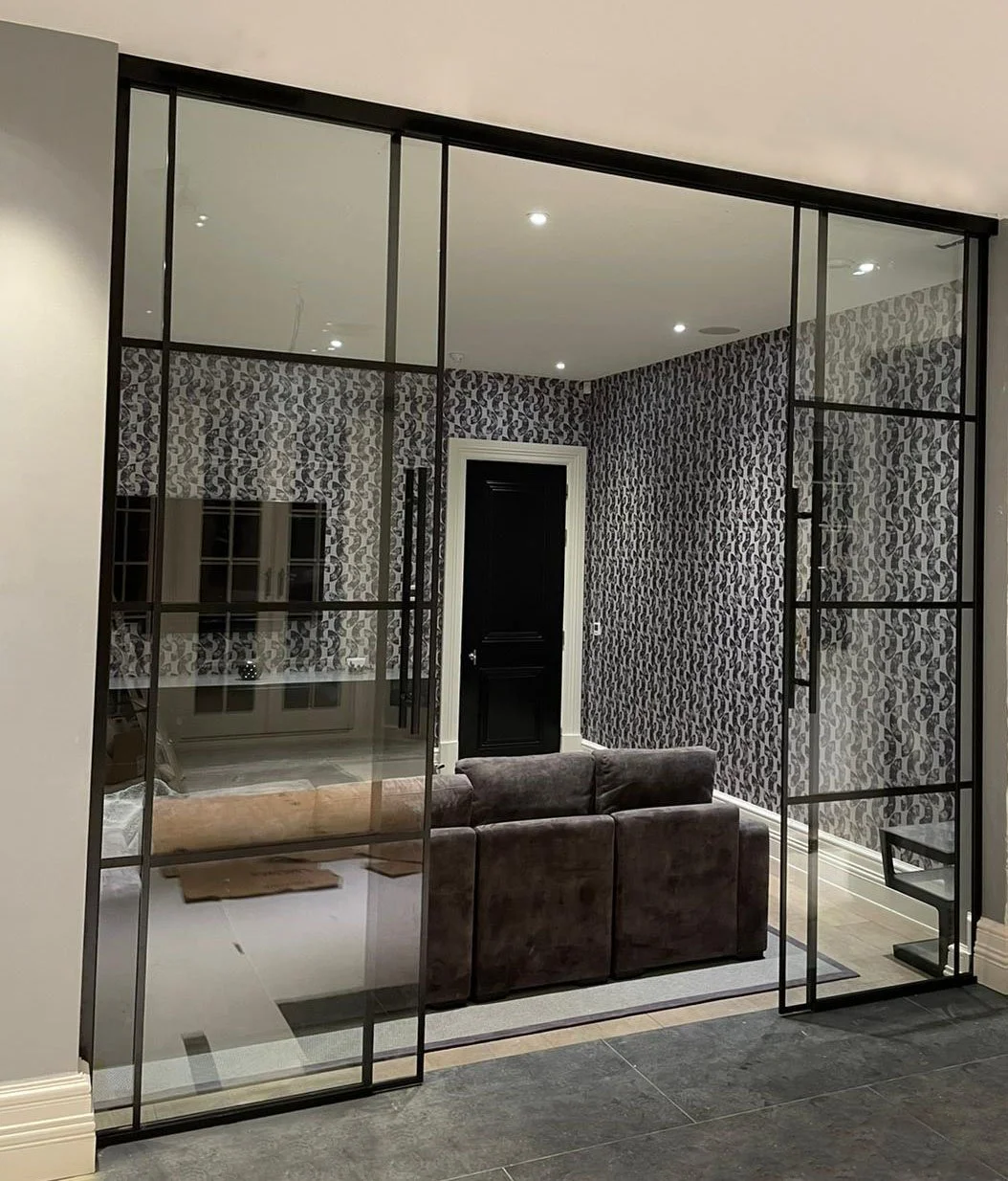 Living room viewed through a large glass sliding door with black metal frames, featuring patterned wallpaper, a black door, a brown sofa, and a pale tiled floor.