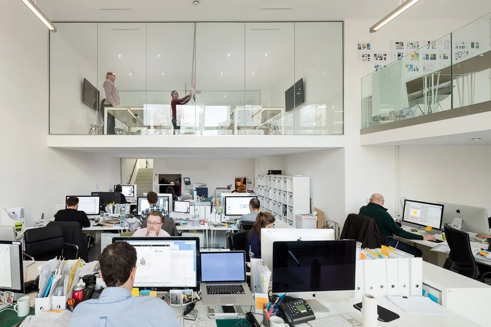 Office with multiple employees working at desks, large white walls, and a glass-walled mezzanine with two people, one using a tablet and another watching.