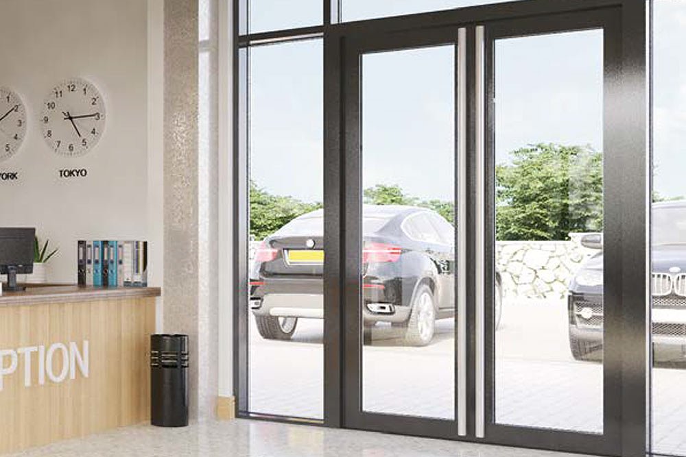 Interior of a modern building’s reception area with a glass door entrance, a wall clock showing around 3:48, a computer on a wooden counter, binders, a small potted plant, and cars visible outside.