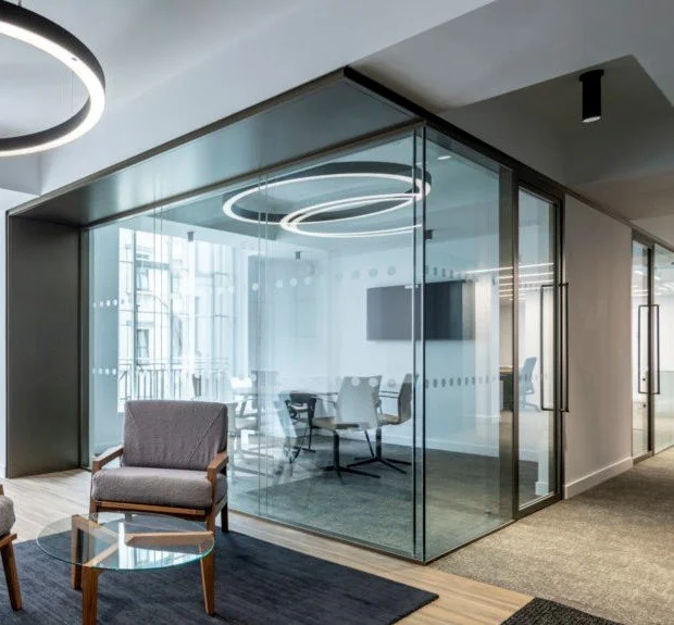 Modern conference room with glass walls, circular ceiling lights, a large TV monitor, and a table with chairs. Front area has upholstered chairs and a small glass side table.