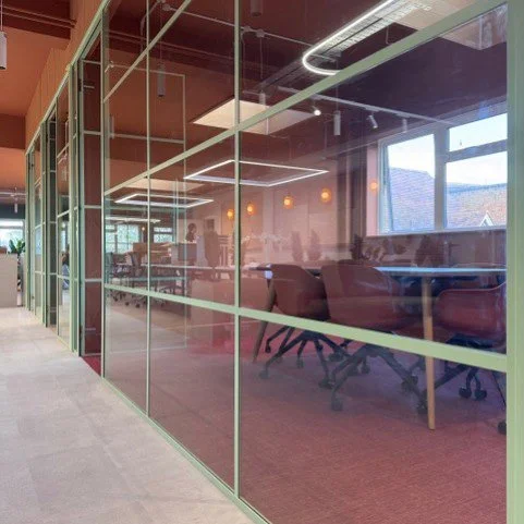 Modern office conference room with pinkish walls, glass walls with white frames, and round pendant lights, seen through glass partitions.