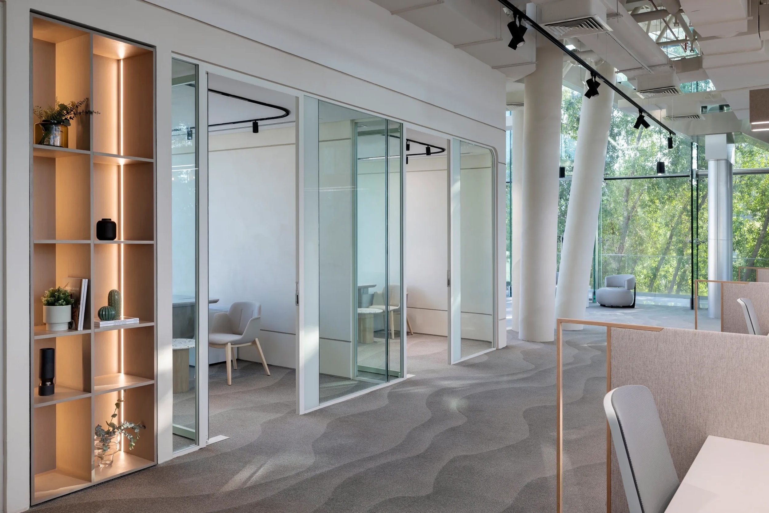 Modern office interior with glass partition walls, a tall wooden shelf with decorative items, chairs, and large windows showing green trees outside.