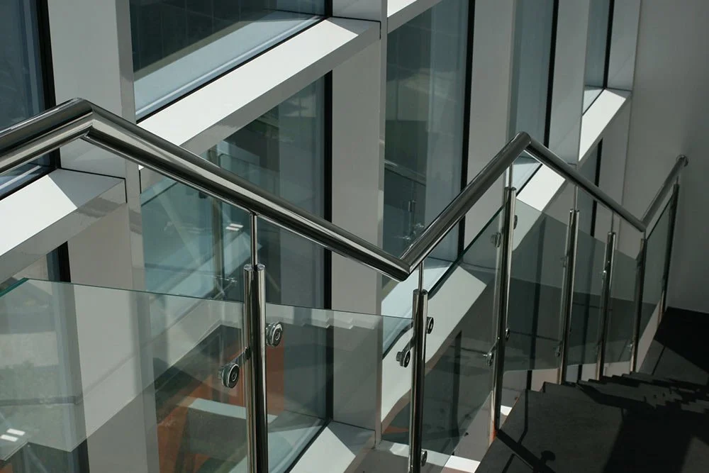 Modern indoor staircase with glass railings and metallic handrails, adjacent to glass walls and windows in a brightly lit building.