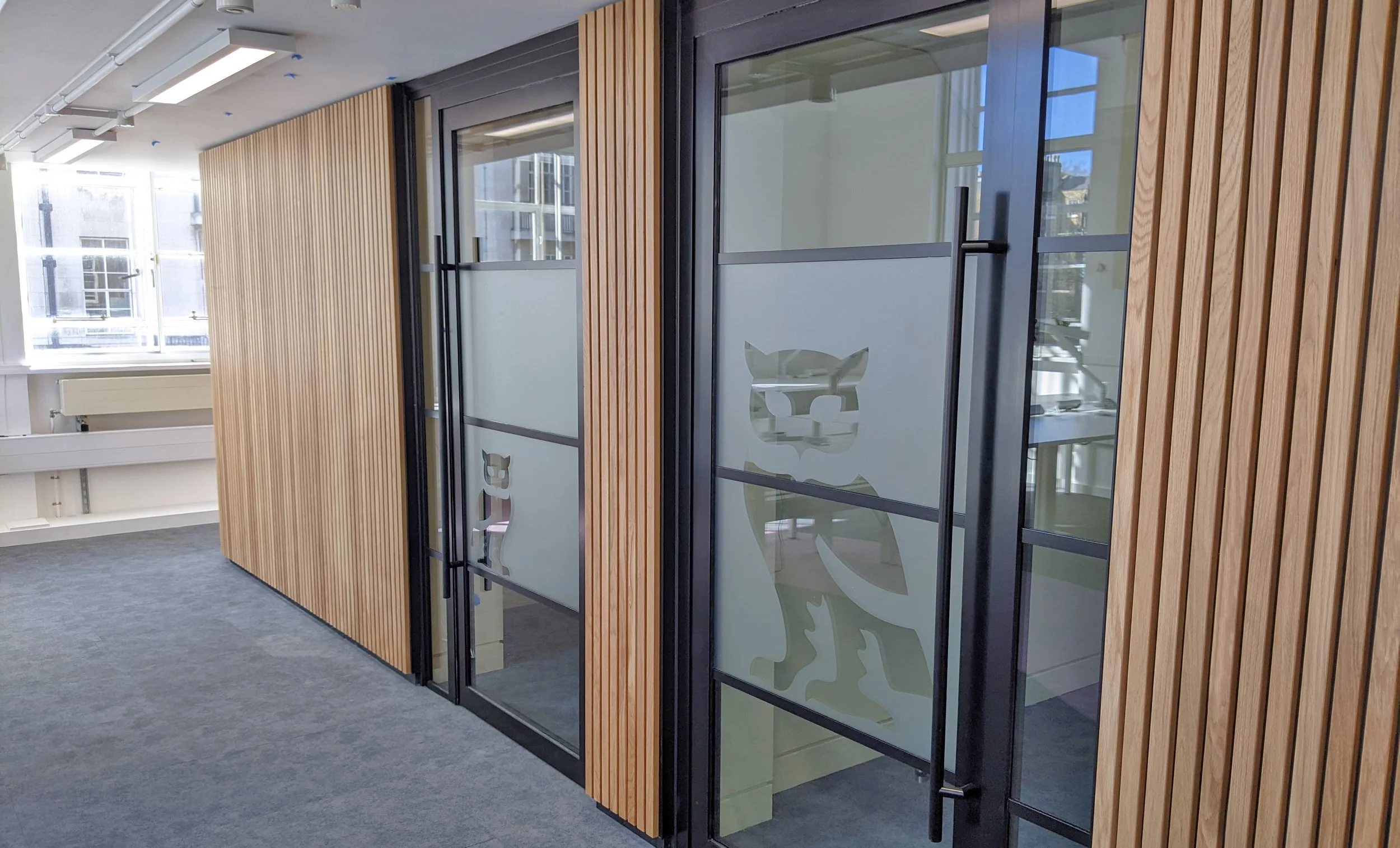 Interior view of office space with wooden wall panels, glass doors with feline logos, and large windows allowing natural light.