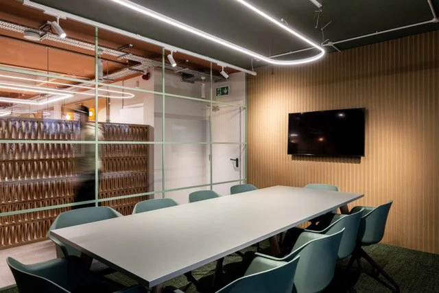 Modern conference room with a long white table, eight blue chairs, a flat-screen TV on a wooden wall, and glass walls separating it from another room.