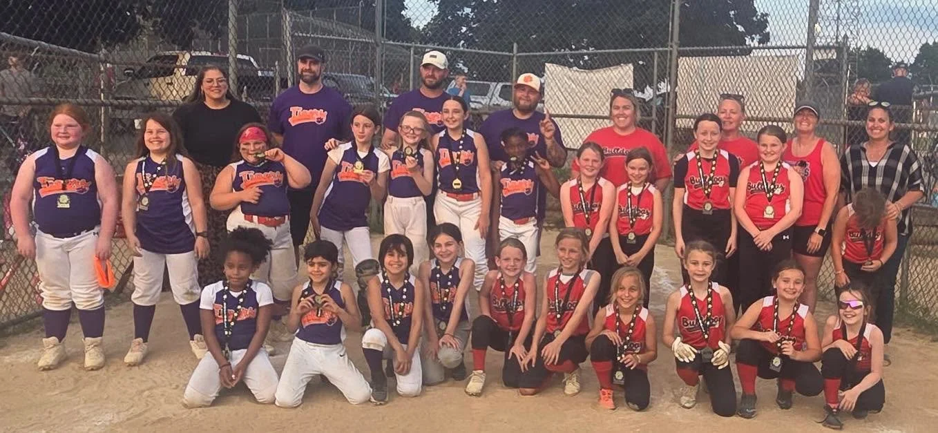 Group photo of young girls and adult women wearing softball uniforms with medals, posing on a softball field through a chain-link fence.