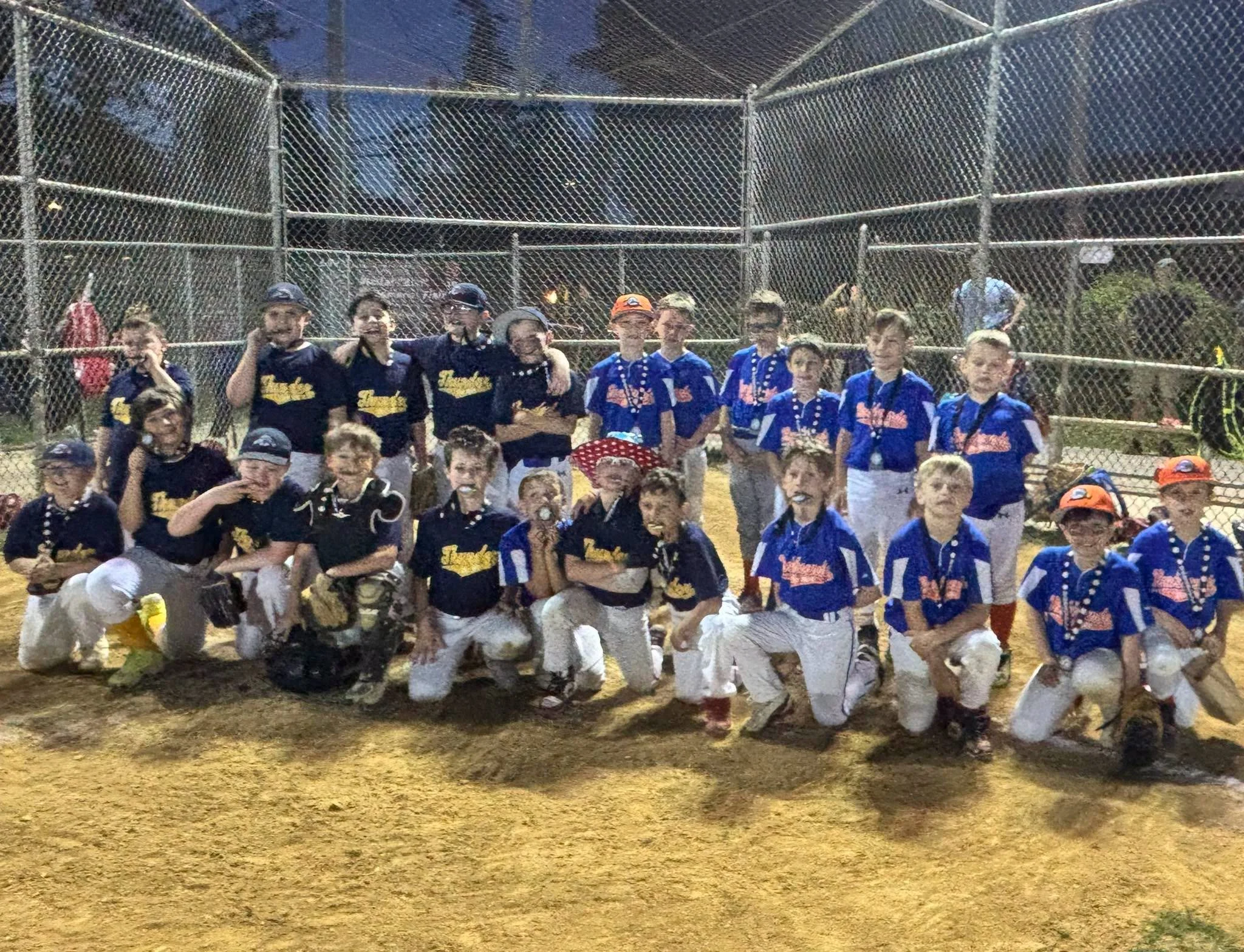 A group of children in baseball uniforms posing together at night on a baseball field, with a chain-link fence and trees in the background.