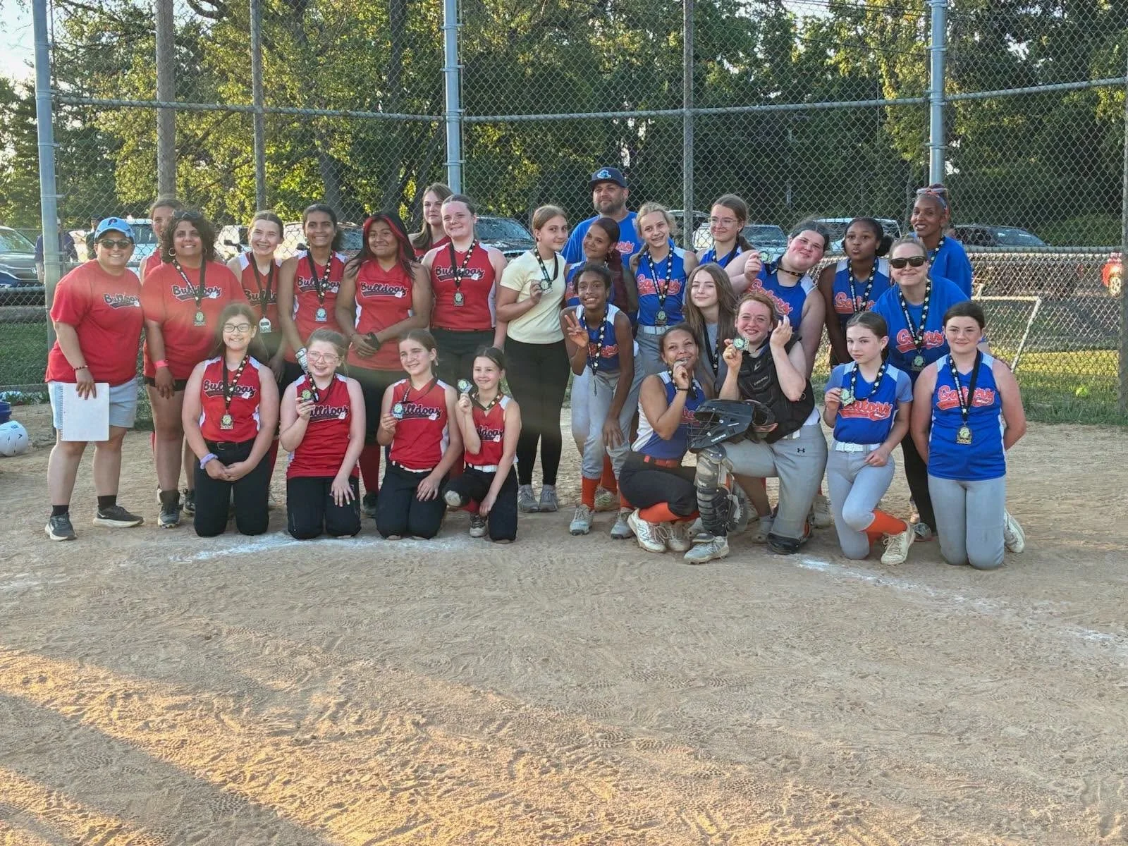 A group of young girls and women on a softball field, celebrating with medals and trophies, some wearing team uniforms and coaches, after a game, with chain-link fences and trees in the background.