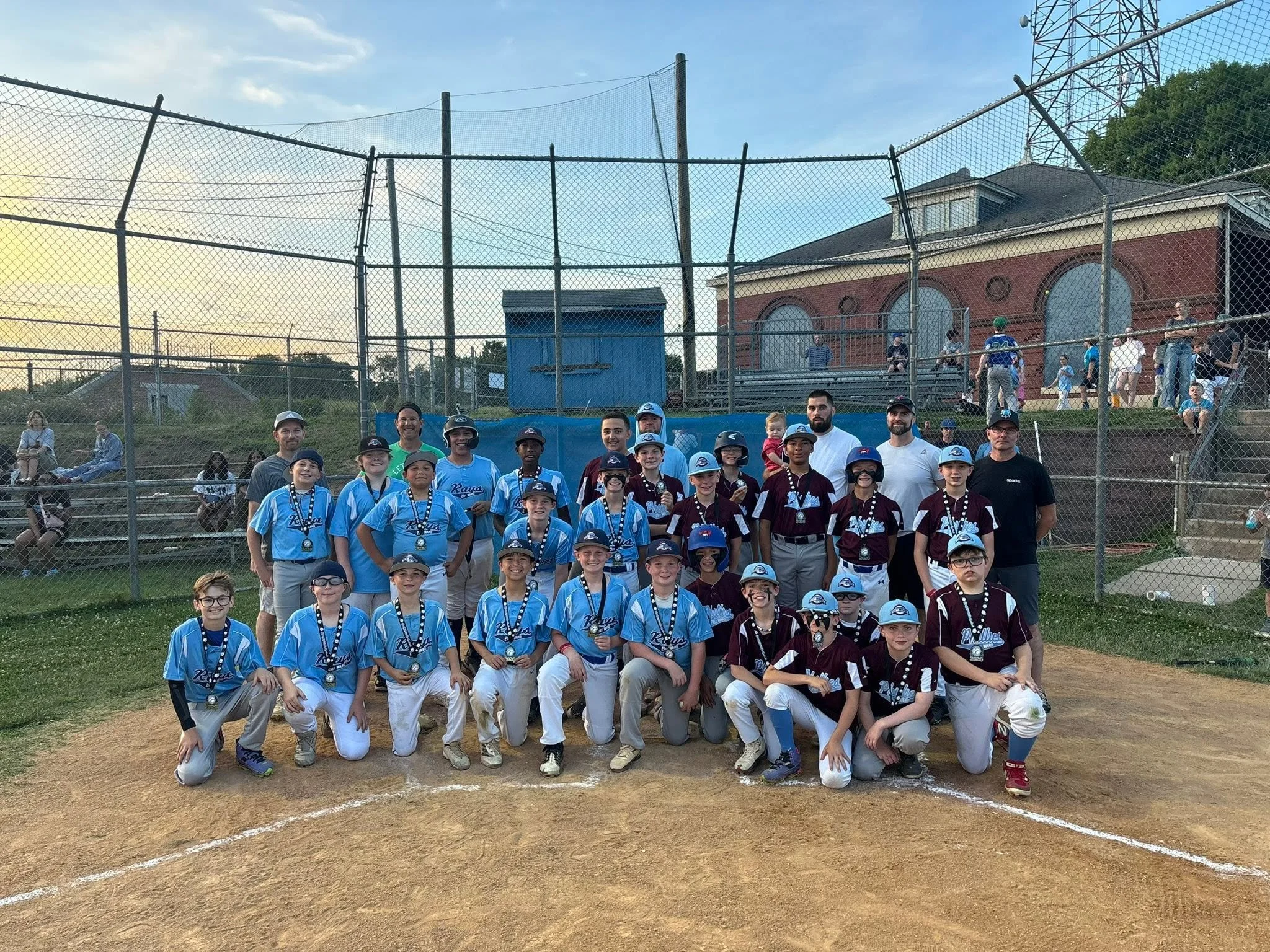 A group of young baseball players and coaches posing for a team photo on the field, with a baseball diamond, fencing, and a building with a tower in the background.