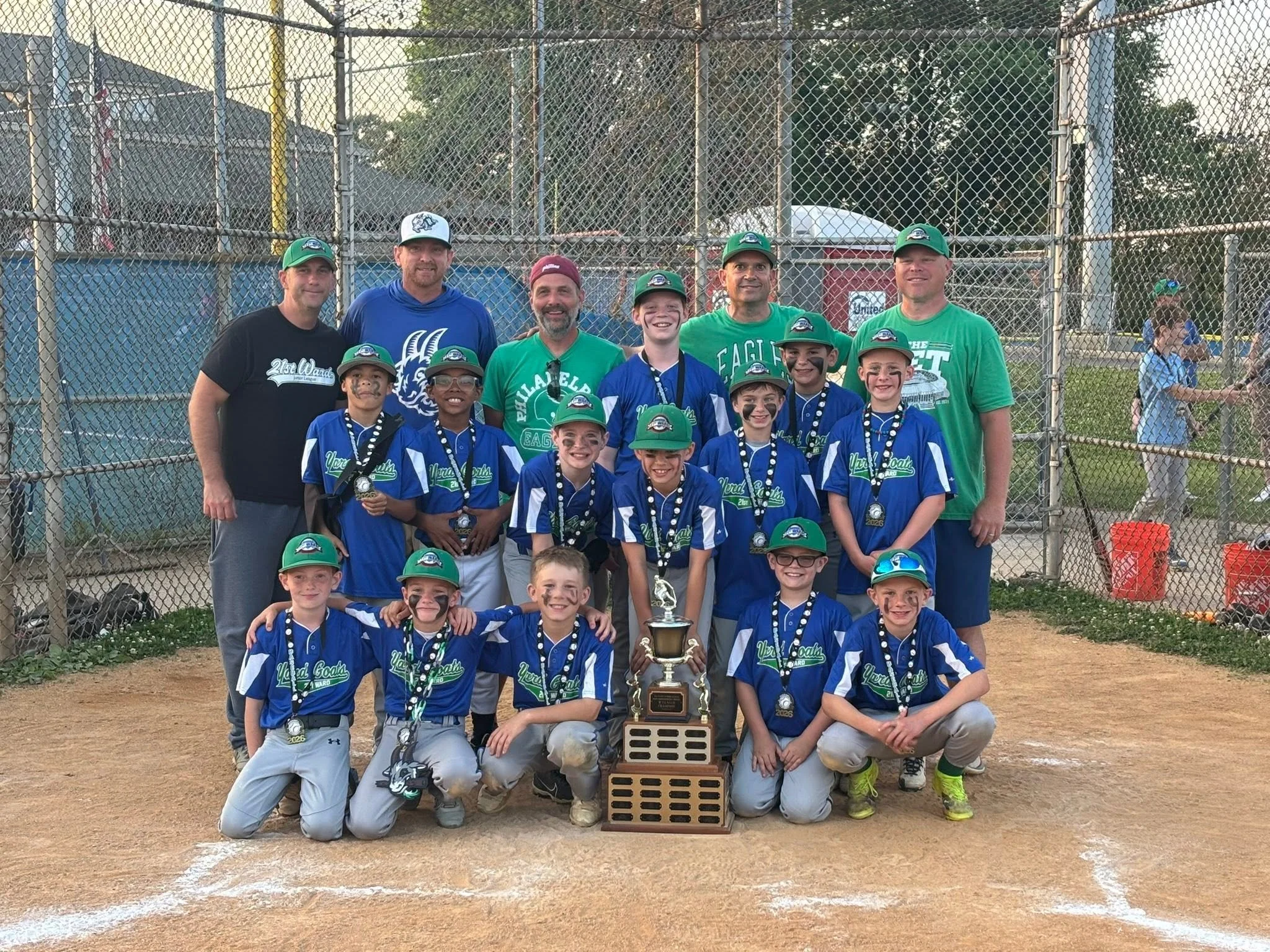 A youth baseball team with coaches and children, holding medals and a trophy, posing on the baseball field after winning a game.
