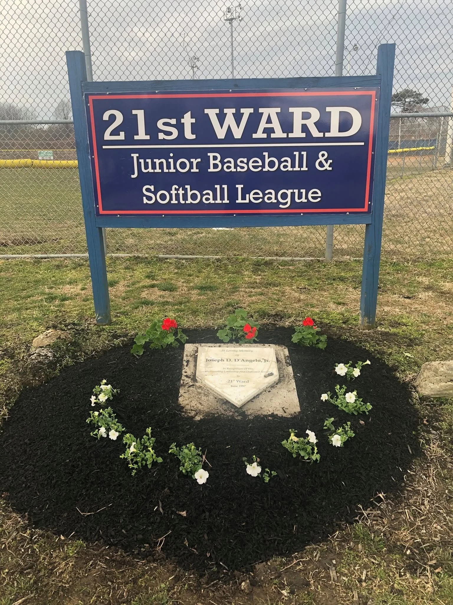 A grave with a white headstone surrounded by flowers, located in front of a blue sign post that reads, "21st WARD Junior Baseball & Softball League." The background features a chain link fence with fields and trees beyond.