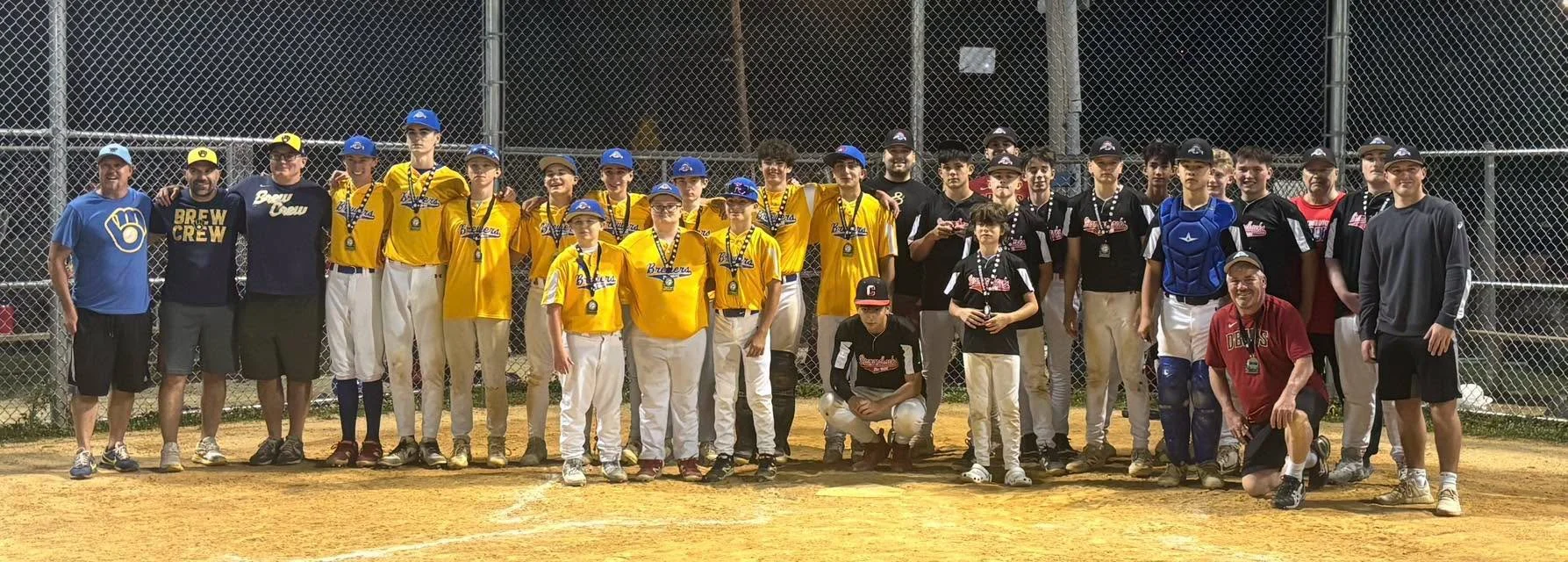 Group of young baseball players and coaches on a baseball field at night. Some players wear yellow jerseys, others wear black jerseys, and some wear catcher's gear. They are posing in front of a chain-link fence.