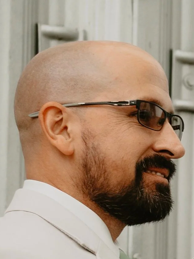 Profile side view of a bald man with a beard and glasses, wearing a white shirt, standing in front of a light-colored wooden wall.
