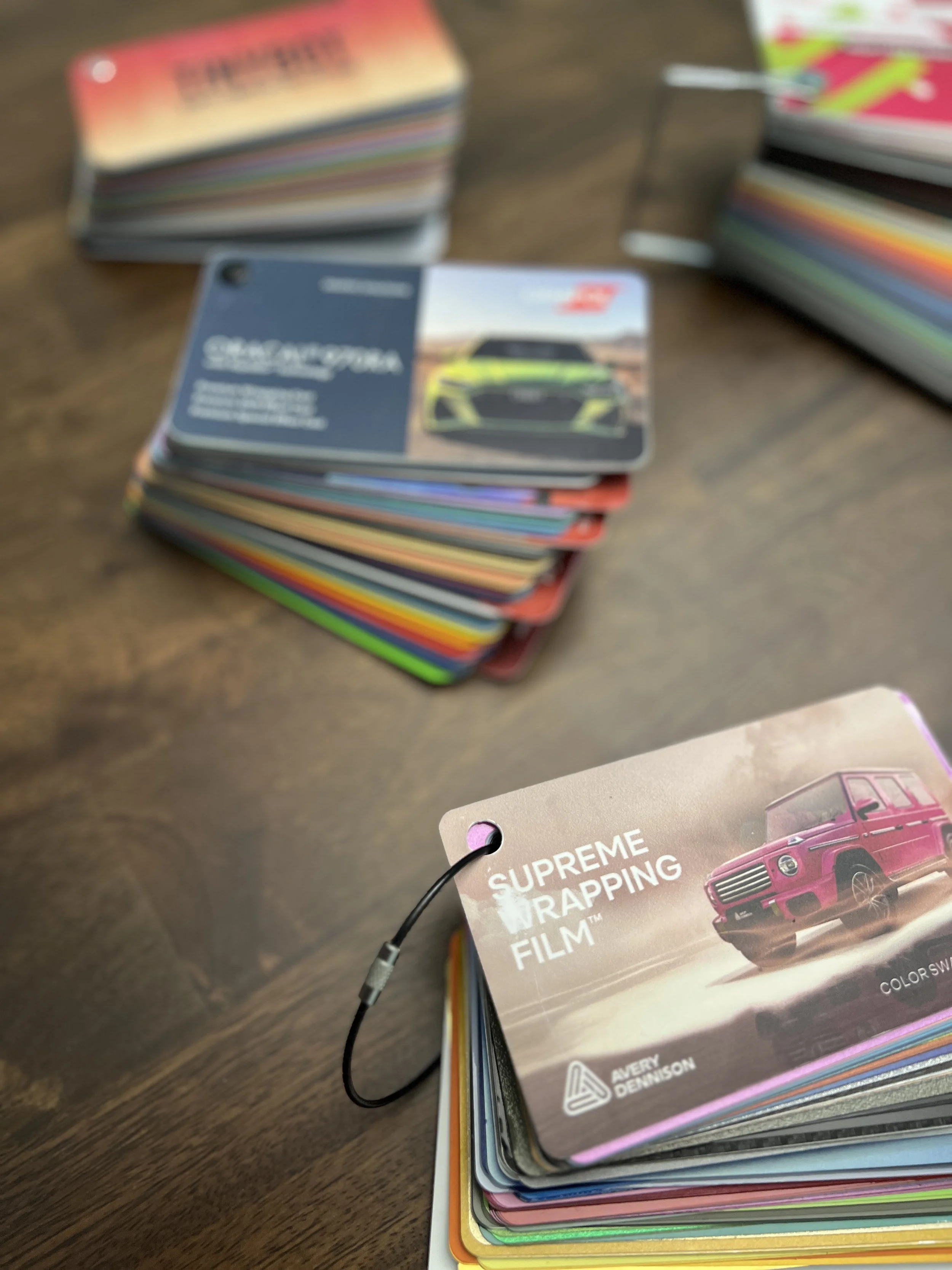Stacked trading cards on a wooden surface, with a visible card featuring a green and black car and a partial card showing a pink vehicle and the text "Supreme Wrapping Film."