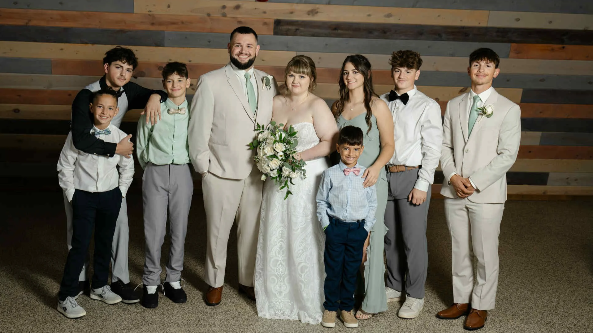 A group of nine people, including adults and children, dressed in formal attire, posing for a photo indoors against a wooden wall.