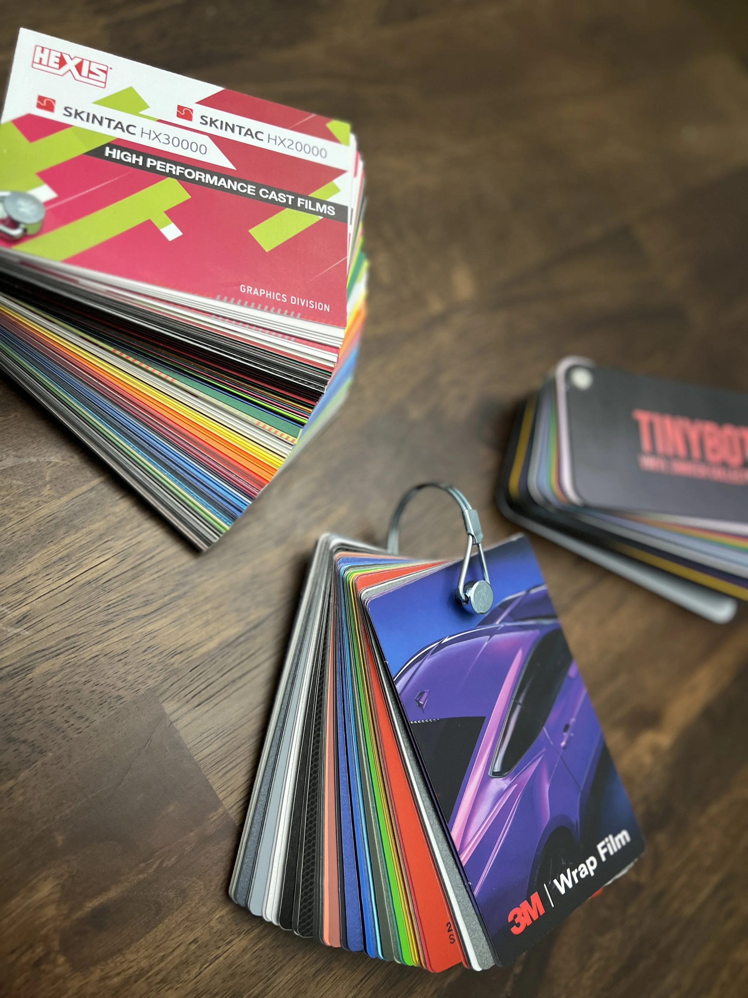 A collection of color sample cards for 3M automotive wrap films, fanned out on a wooden surface. The top card displays the 3M logo, product name, and information about high performance cast films.