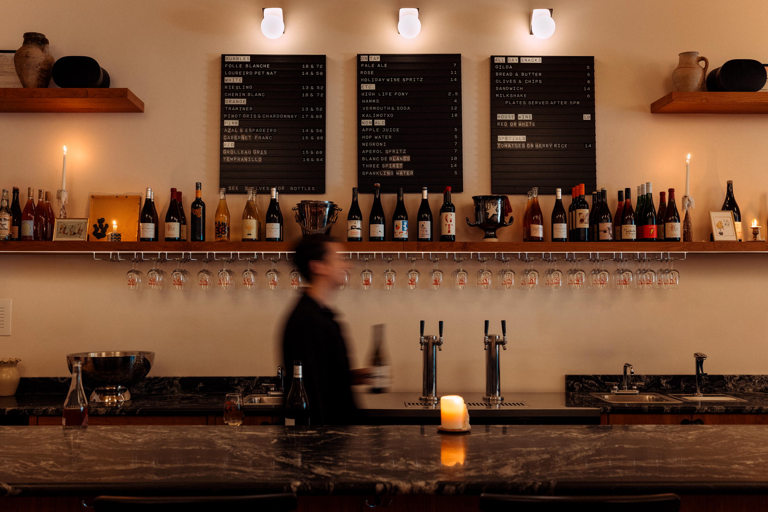 A bar with a dark granite counter, featuring a candle, and a row of various wine bottles on a wooden shelf. Two black speaker containers and two earthenware vases are mounted on the wall above. The wall displays multiple blackboards with handwritten menus listing drinks and snacks. A blurred figure is seen in motion, likely a bartender.
