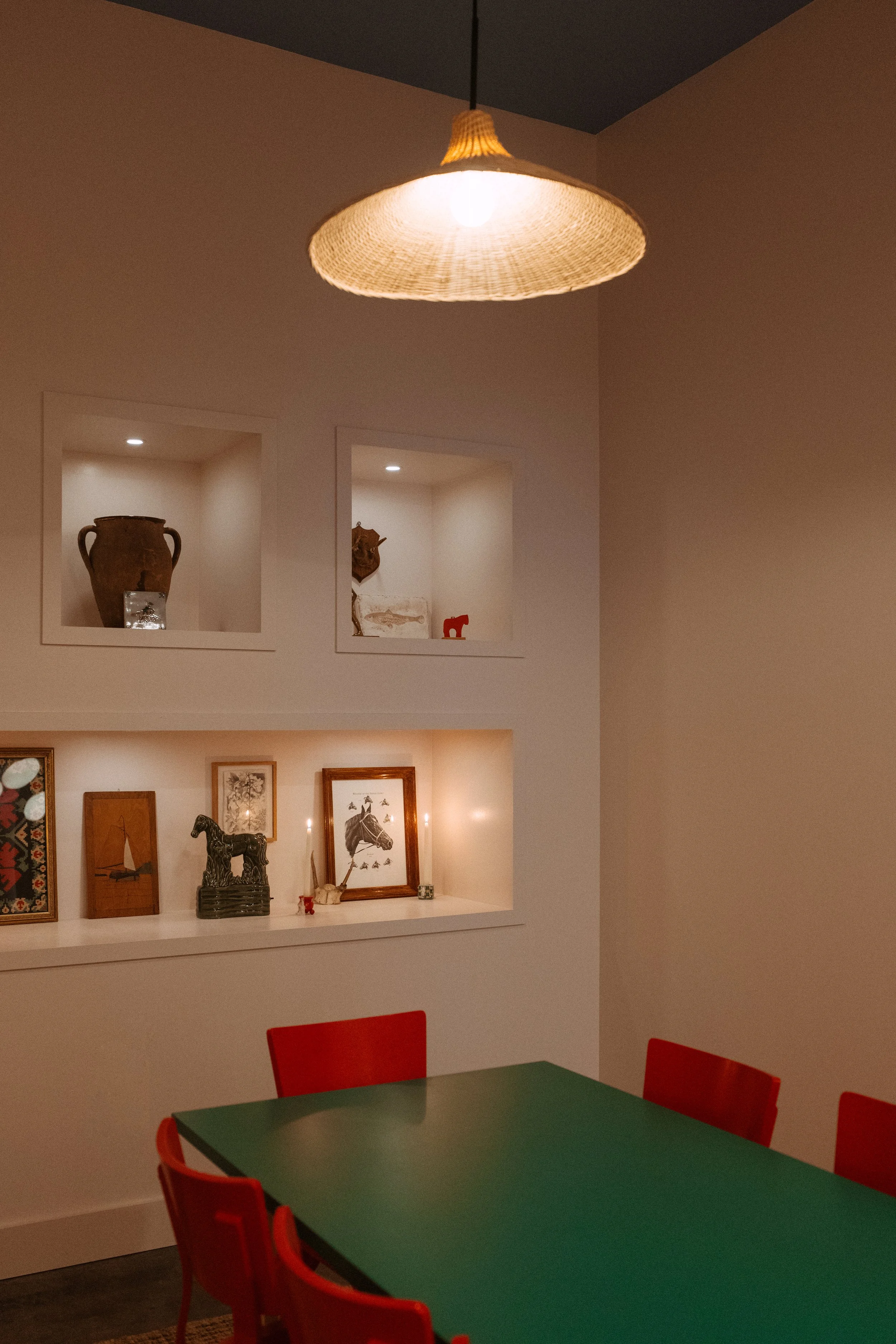 A room with a green table and red chairs, two shadowed display shelves containing decorative items such as pottery and framed art, and a hanging ceiling light with a woven shade.