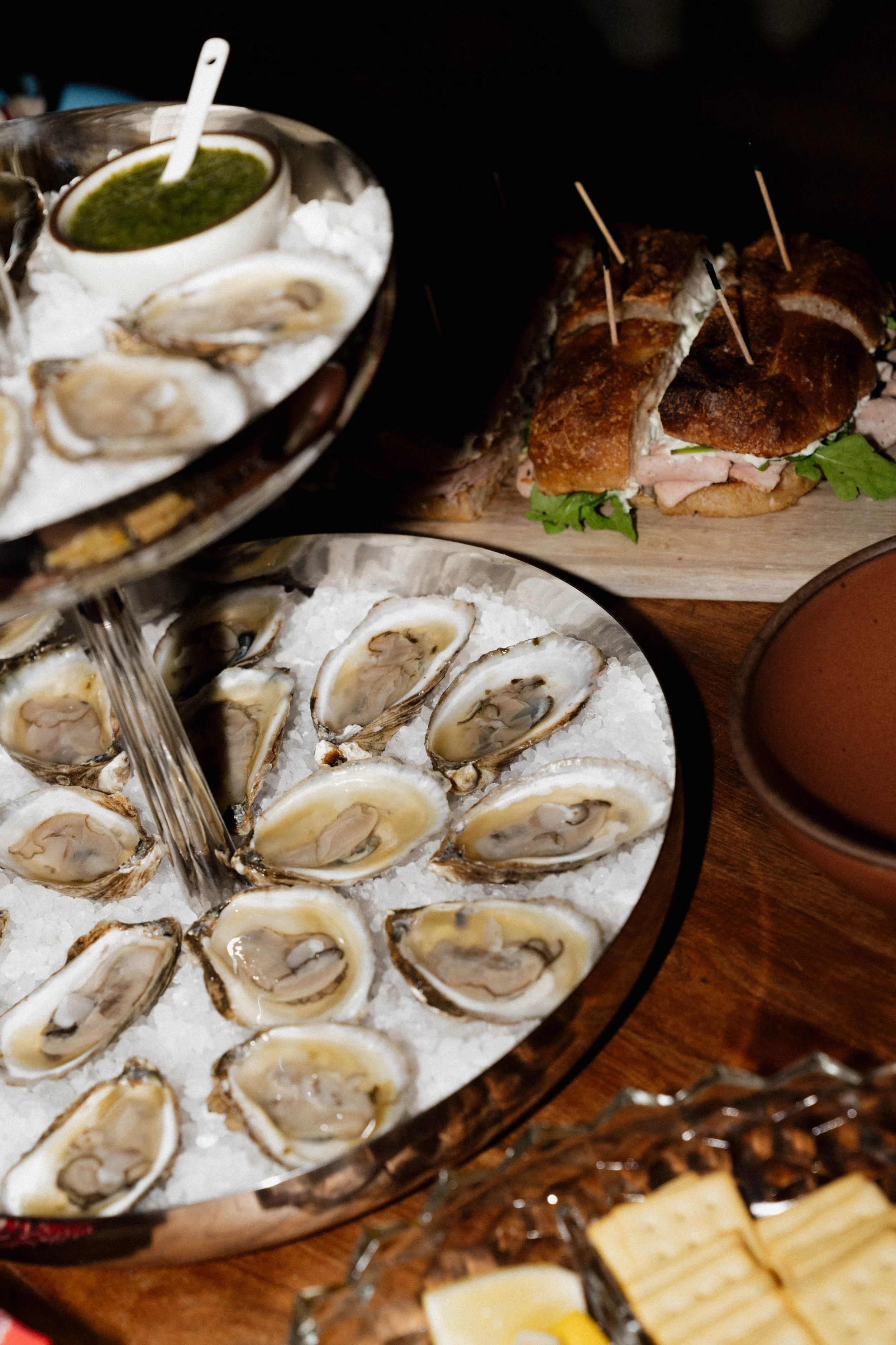A seafood platter with fresh oysters on ice in the foreground and a sandwich with meat, greens, and toothpicks in the background.