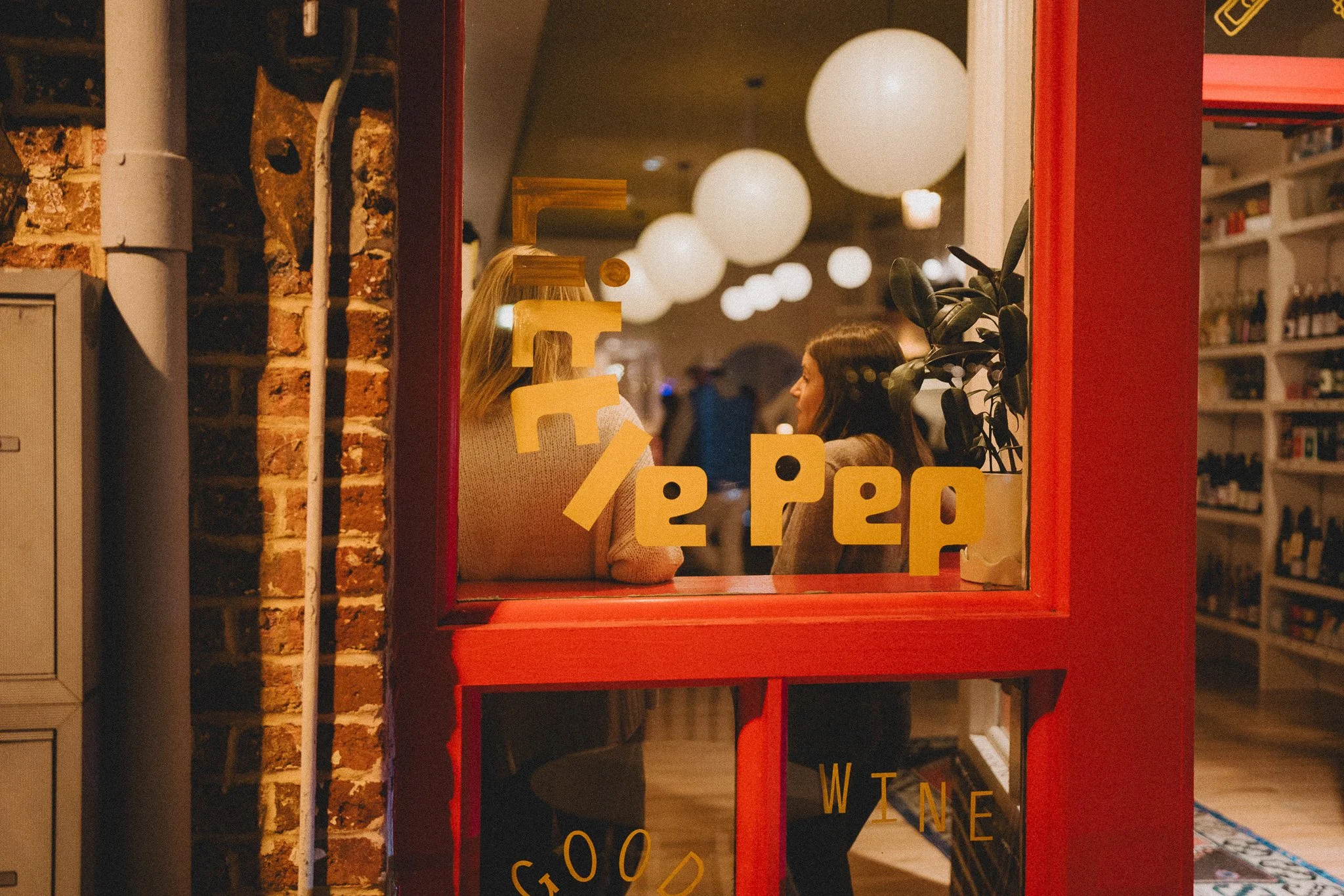 Looking through a window in a restaurant or café, two women are seated at a table, engaged in conversation. The window has yellow letters and words on it, and inside, hanging spherical lights illuminate the space. The interior features exposed brick walls and shelves with various items.