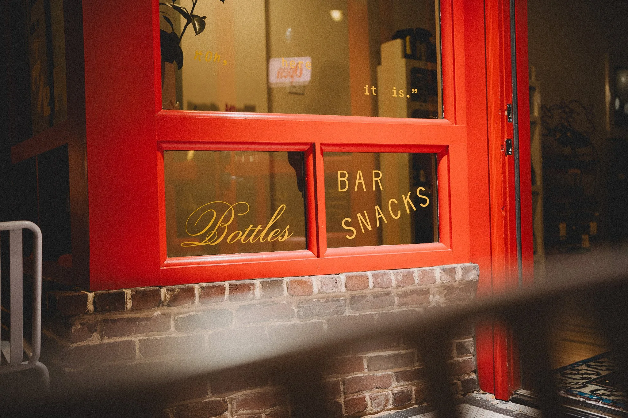 Red-painted window at a bar or snack shop with the words 'Bottles,' 'Bar,' and 'Snacks' on the glass. Inside, a reflection of a sign reads 'here.' The building has a brick base, and there is a small mat with a colorful pattern outside.