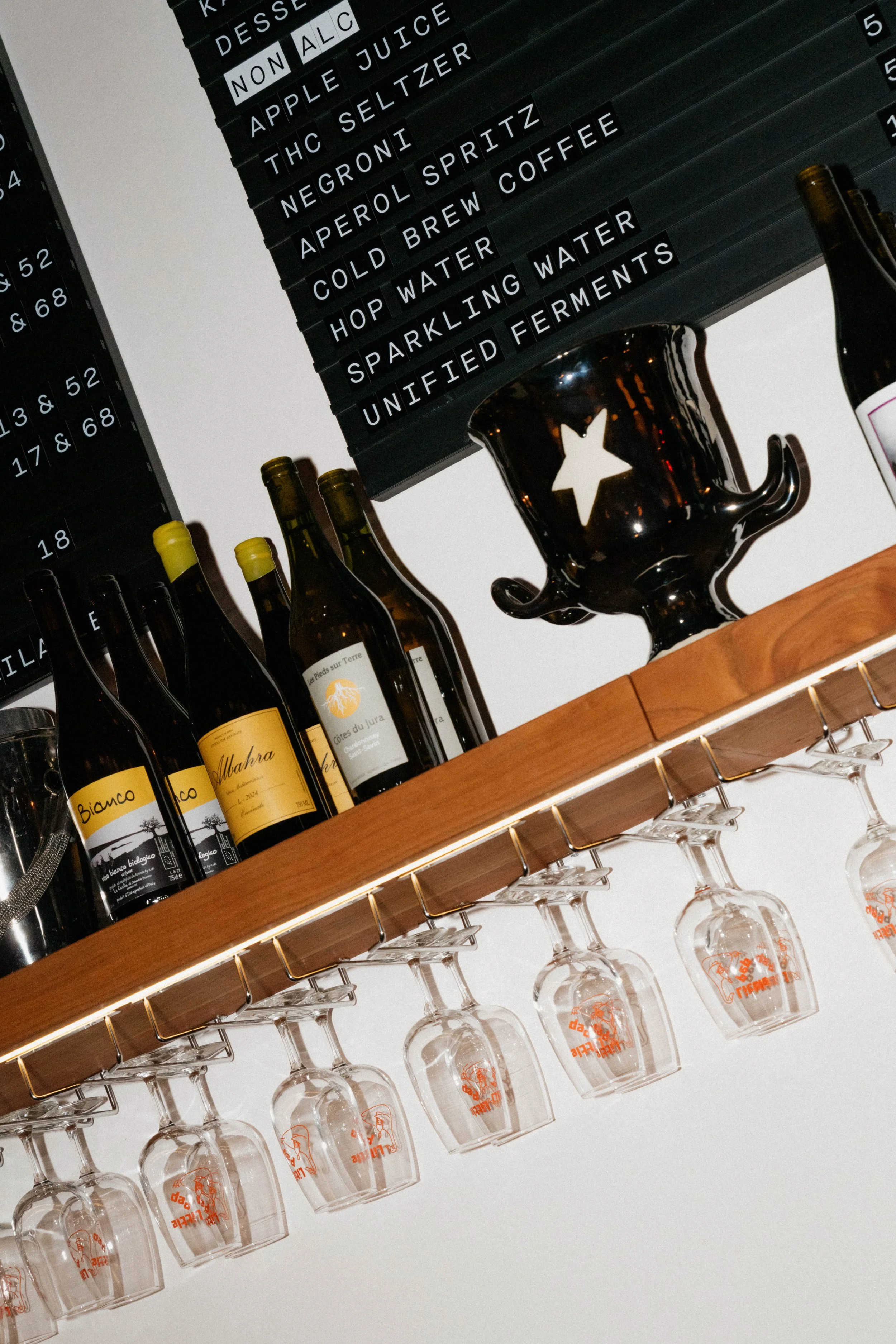 A bar or kitchen shelf displaying wine bottles and wine glasses hanging upside down. A black board with white text lists drink options, including non-alcoholic beverages, hard seltzer, juice, and fermented drinks.