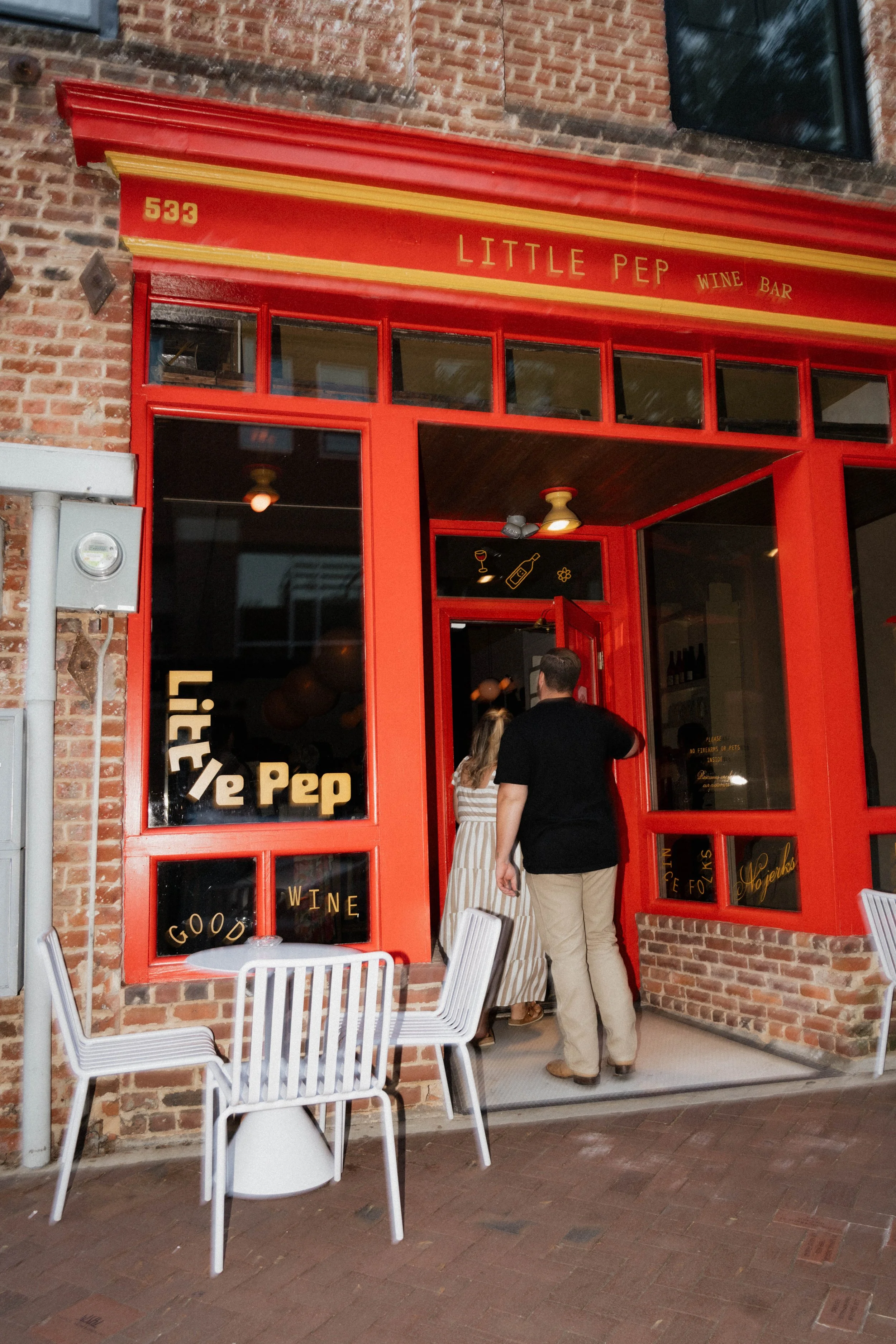 People entering Little Pep Wine Bar through a red brick storefront with large windows. Outside seating with white chairs and tables, and a blackboard sign.