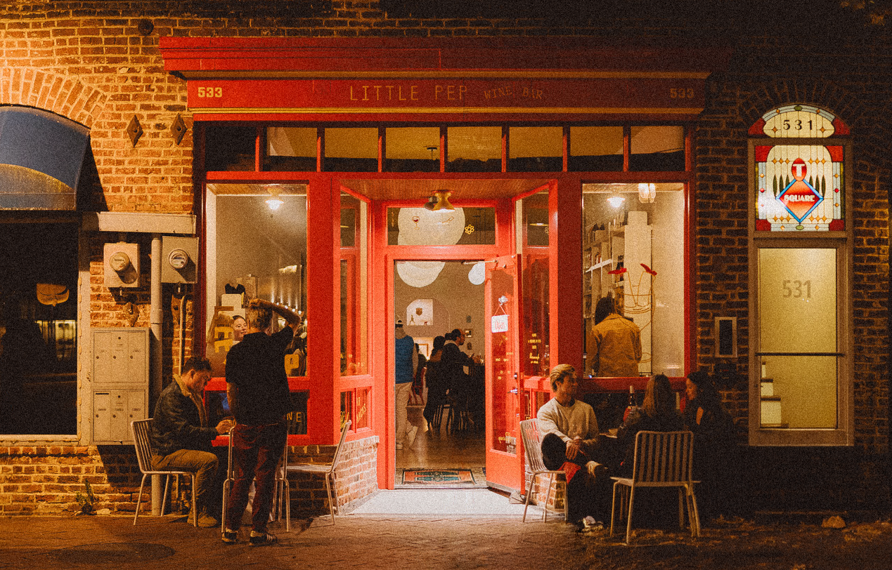 Nighttime scene of a brick building with a bright red entrance labeled 'Little Pep Wine Bar'. Several people are sitting outside, some engaged in conversations and one using a phone, while a person standing by the door is pouring a drink. The interior is visible through the glass windows and open door.