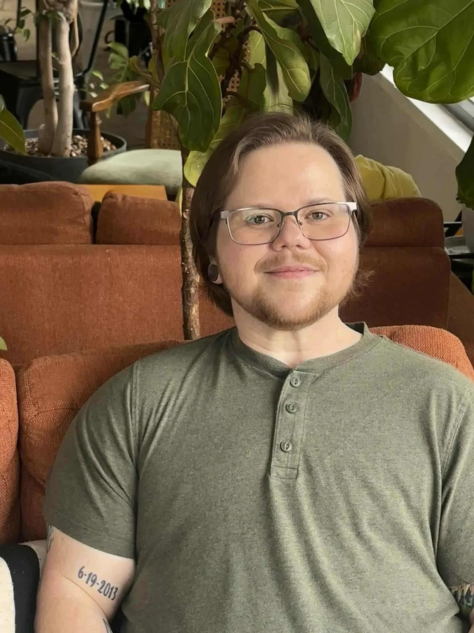A masculine perceived human with glasses and facial hair wearing a light green henley shirt, sitting on a rust-colored sofa with green plants in the background, their arm with a tattoo of the date '6-19-2013' is visible.