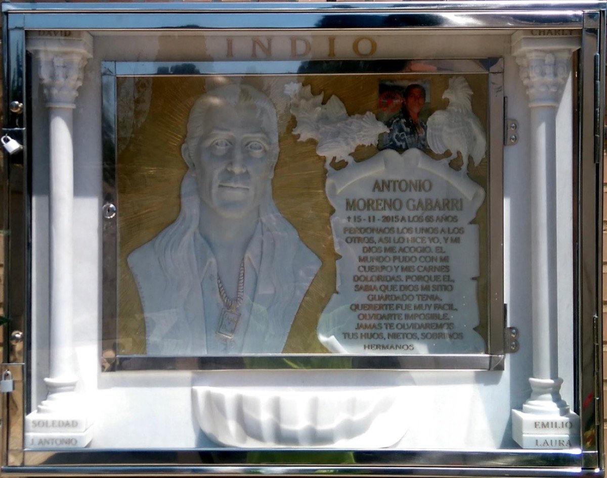 Impresionante lápida escultórica en mármol blanco de Carrara, protagonizada por el busto tallado del difunto con detalles minuciosos como su cadena, realzada en auténtico pan de oro al igual que todo el fondo de la obra. La composición integra dos ga