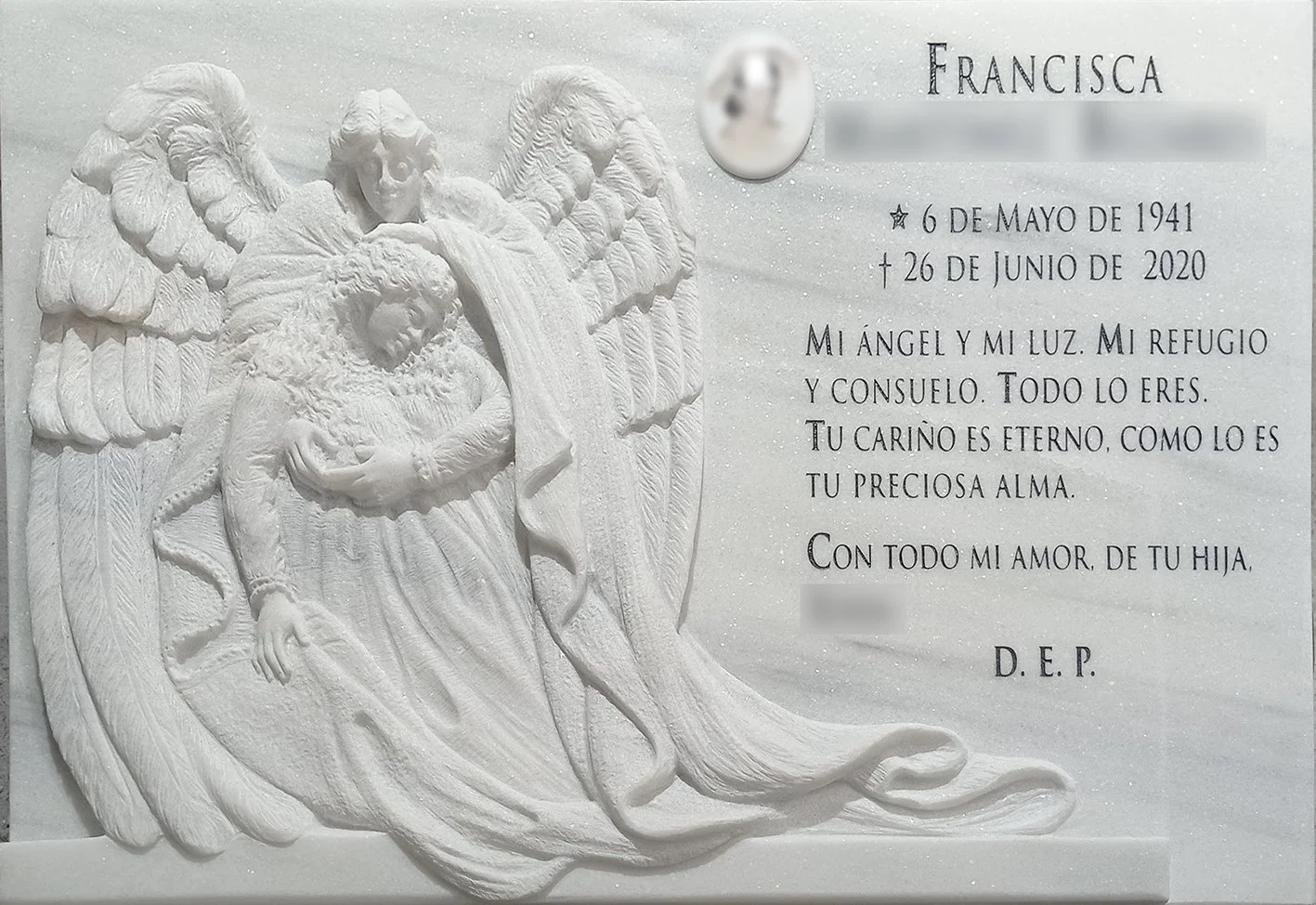 Lápida en mármol blanco tallada a mano con la figura de un ángel y la virgen en relieve rebajado, destacando por el minuciosos trabajo de detalle en las plumas de las alas y los pliegues de los mantos. La composición se completa con una elegante insc