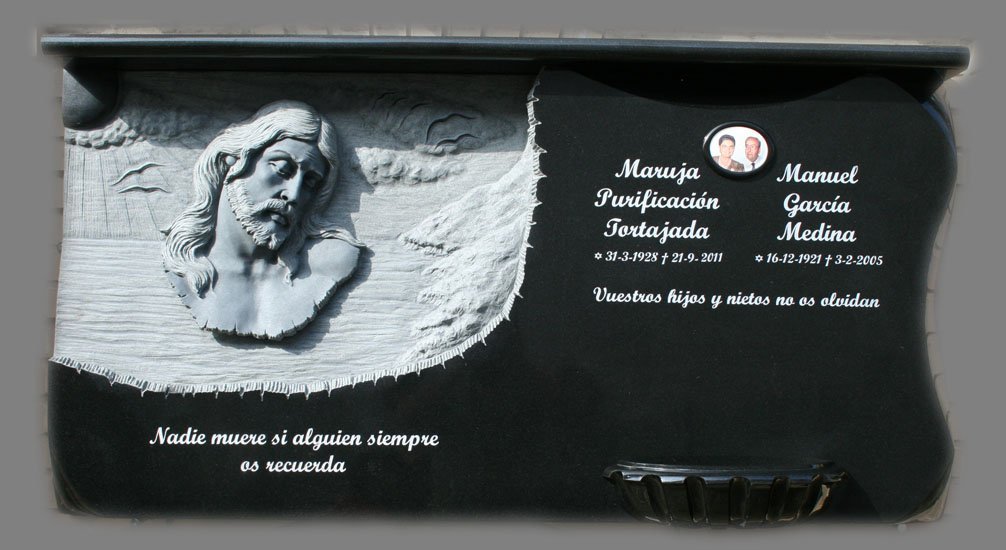 Lápida esculpida en granito negro, protagonizada por una detallada cabeza de Cristo acompañada de ornamentación en la parte trasera y una elegante jardinera en forma de concha. El diseño se completa con distintivos biseles en el lateral derecho que a