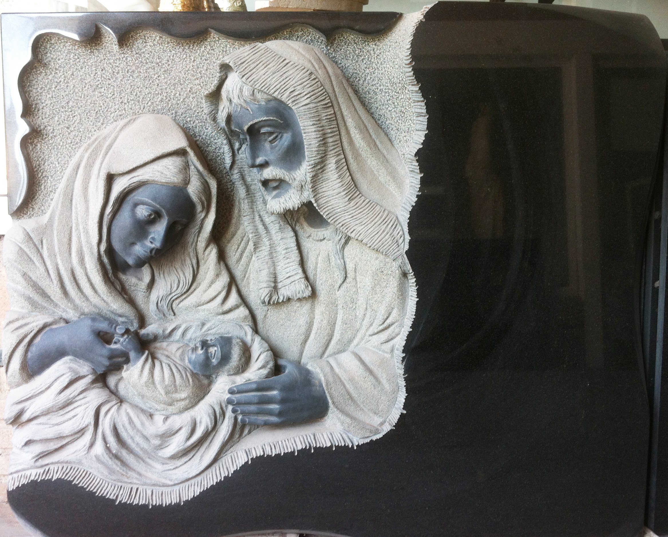 Lápida en granito negro con relieve escultórico de una maternidad enmarcada por un delicado adorno exterior. Obra tallada a mano por el escultor Raúl Abad en nuestro taller de Alfara del Patriarca (Valencia).