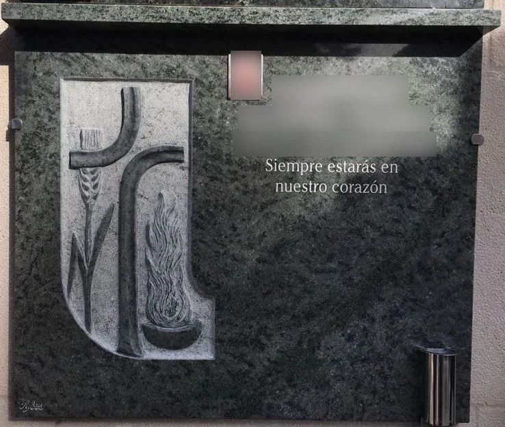Lápida en piedra de granito verde caracterizada por una cruz esculpida que incorpora delicados detalles en relieve de una espiga y una llama, combinada con un moderno búcaro en acero inoxidable. Una pieza de marcada carga simbólica y estética equilib