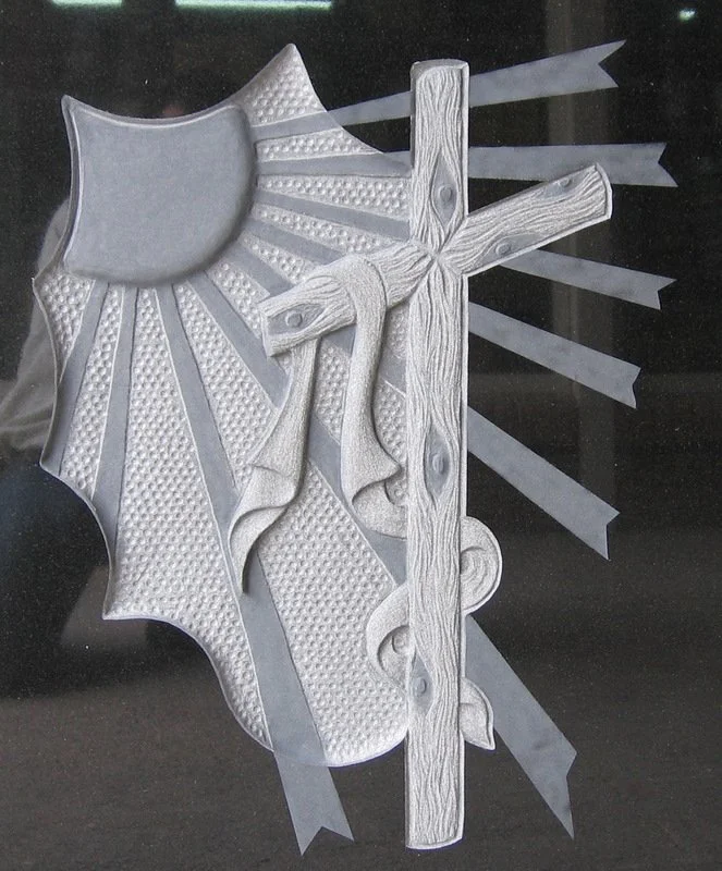 Detalle de escultura tallada a mano con una cruz en bajorrelieve envuelta en un manto, acompañada en la esquina por un sol con sus rayos y un fino fondo repicado artesanalmente. Una delicada muestra de escultura y arte funerario elaborada en nuestro 