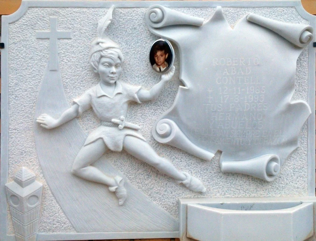 Lápida en mármol blanco Carrara con diseño infantil de Peter Pan, una cruz que da paso a un camino, pergamino con volutas en gran relieve para la inscripción y jardinera sobrepuesta. Obra tallada a mano por el escultor Raúl Abad en nuestro taller de 