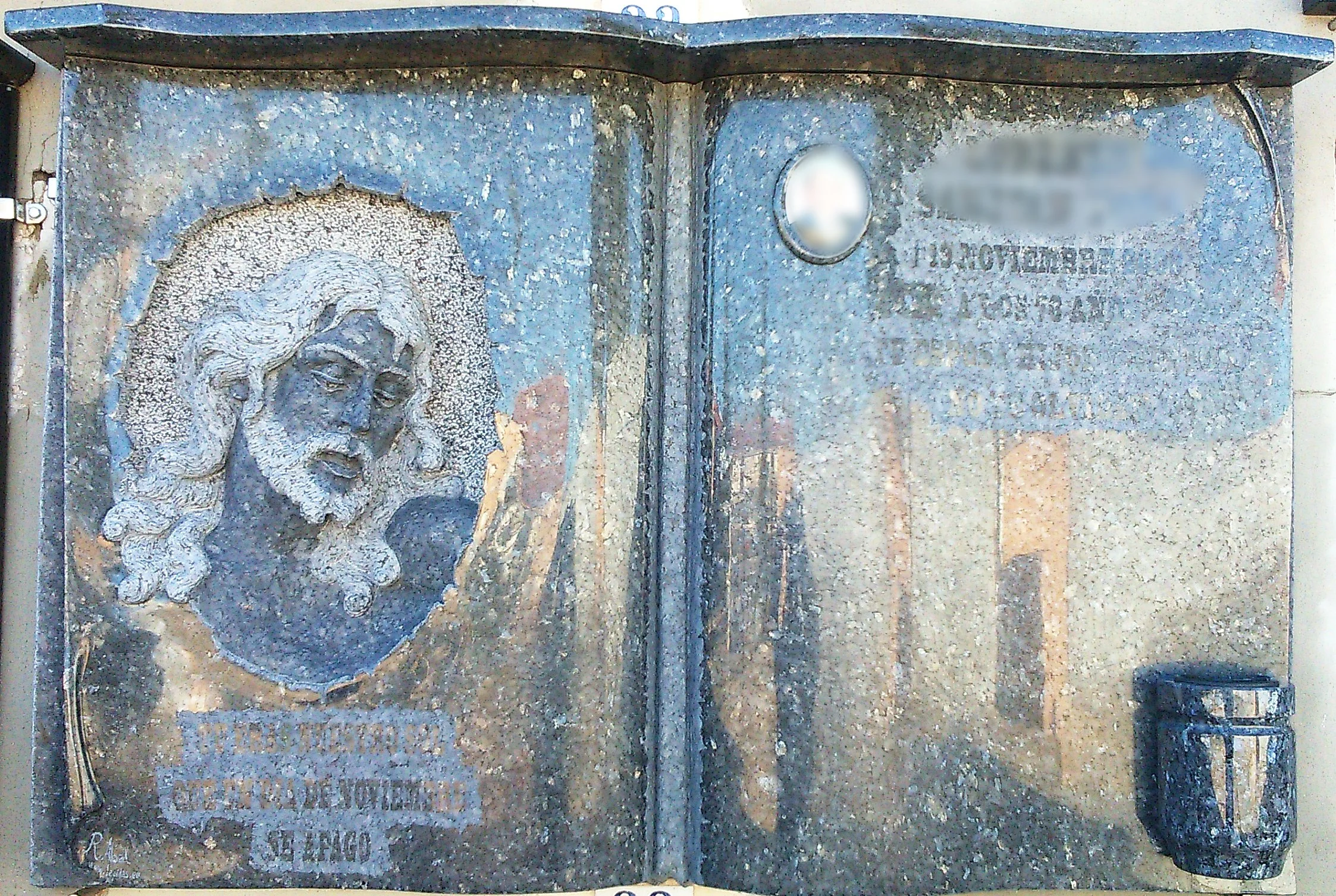 Lápida en granito Labrador Claro tallada en forma de libro abierto, con la escultura en relieve de la cabeza de Cristo sobre una de las páginas, acompañada de inscripción grabada a relieve y búcaro. Obra tallada a mano por el escultor Raúl Abad en nu