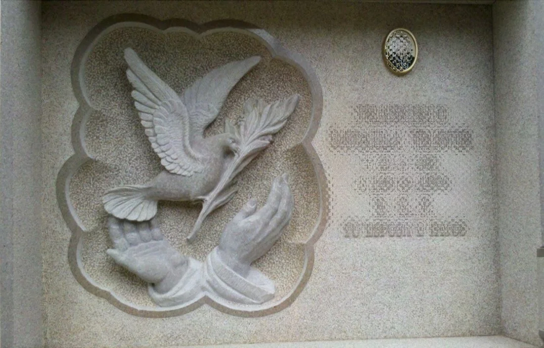 Lápida en mármol de Borriol con escultura en relieve de paloma, manos y rama de olivo. Acabado con superficie repicada y contorno de la paloma e inscripción pulidos. Obra tallada a mano por el escultor Raúl Abad en nuestro taller de Alfara del Patria