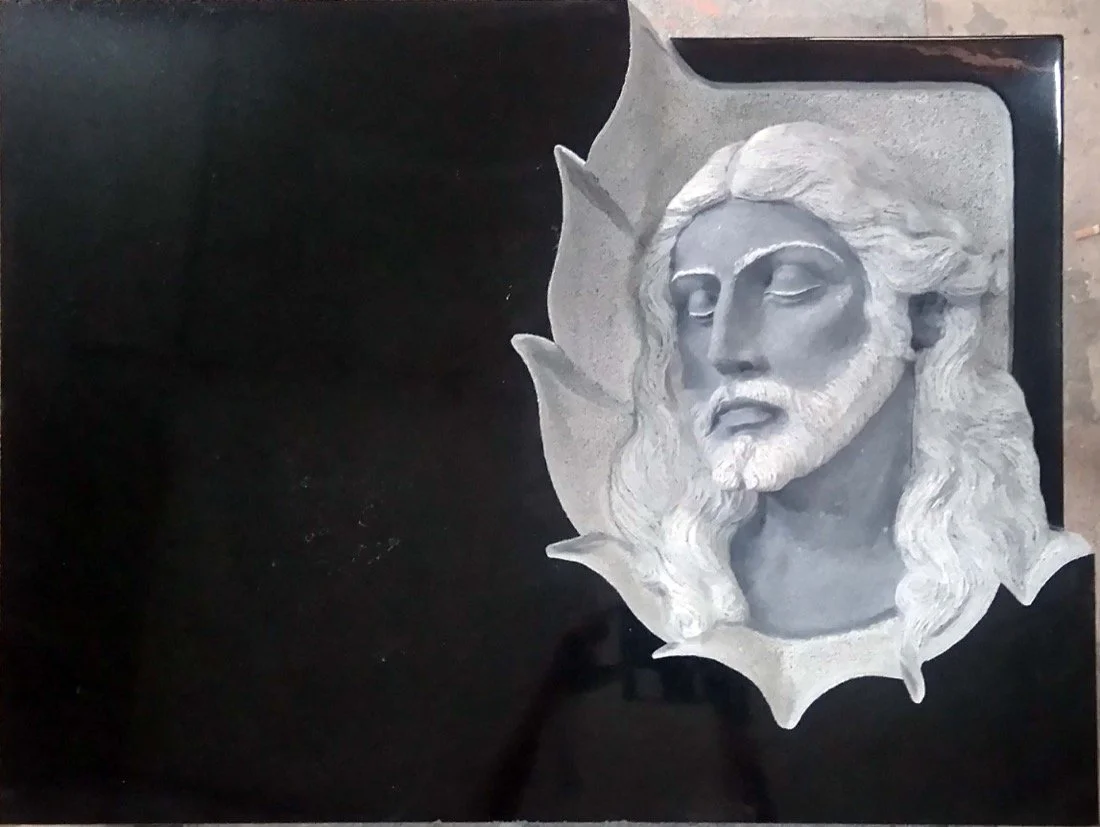 Lápida de granito negro con contorno en forma de hoja y remarcada en la parte derecha abierta, donde se sitúa la escultura en relieve de la cabeza de Cristo. Obra tallada a mano por el escultor Raúl Abad en nuestro taller de Alfara del Patriarca (Val