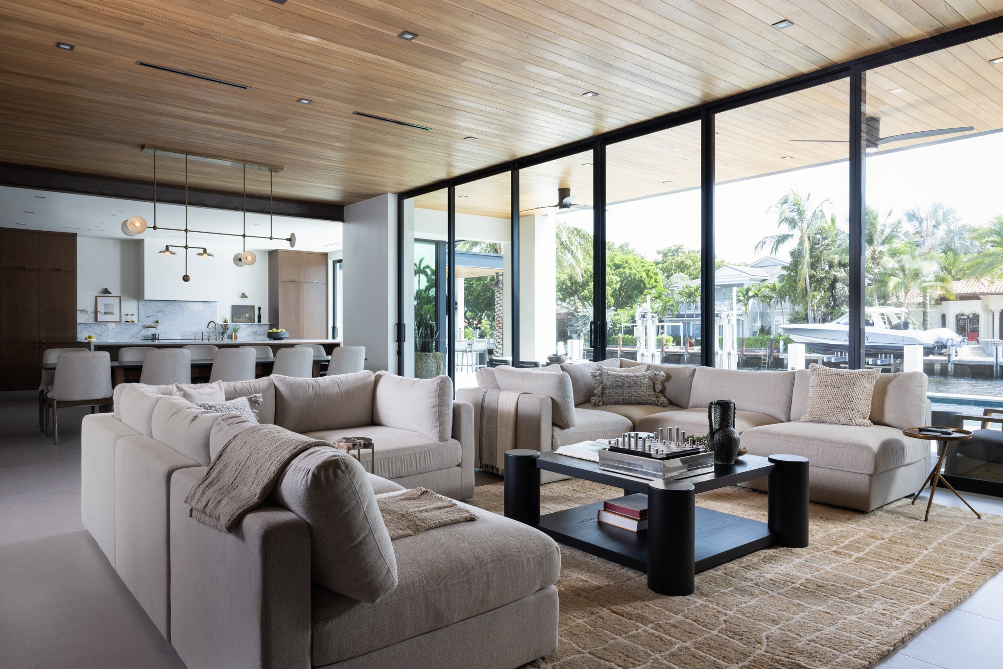 Modern living room with cream interior define sectional sofas, a custom black oak coffee table, and large glass sliders windows and a cedar covered ceiling with linear registers overlooking waterfront homes with boats and palm trees.