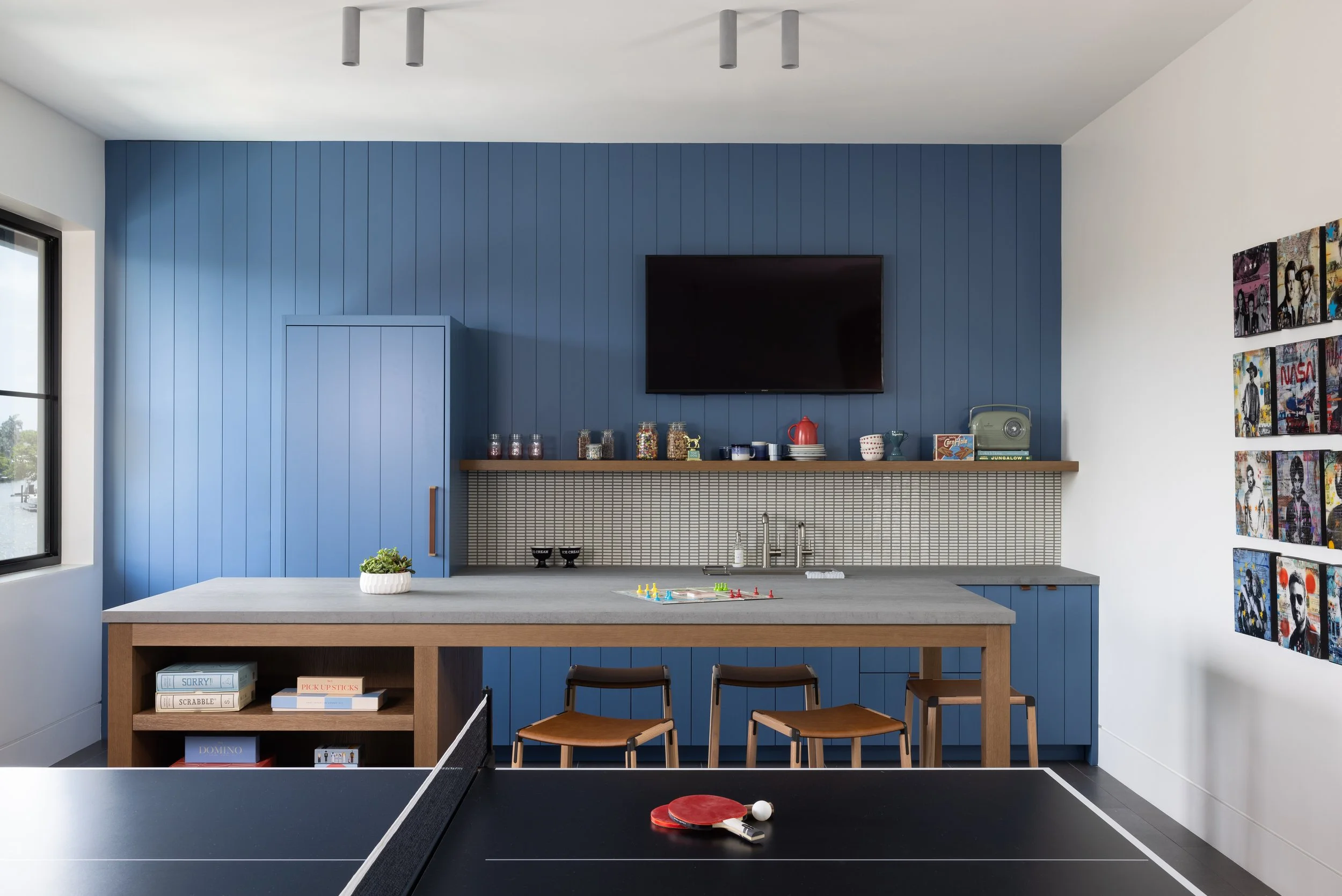 Custom modern blue kitchen with vertical paneled walls and integrated panel-ready appliances, custom artwork and a gray countertop in a teen game room. A custom ping pong table.