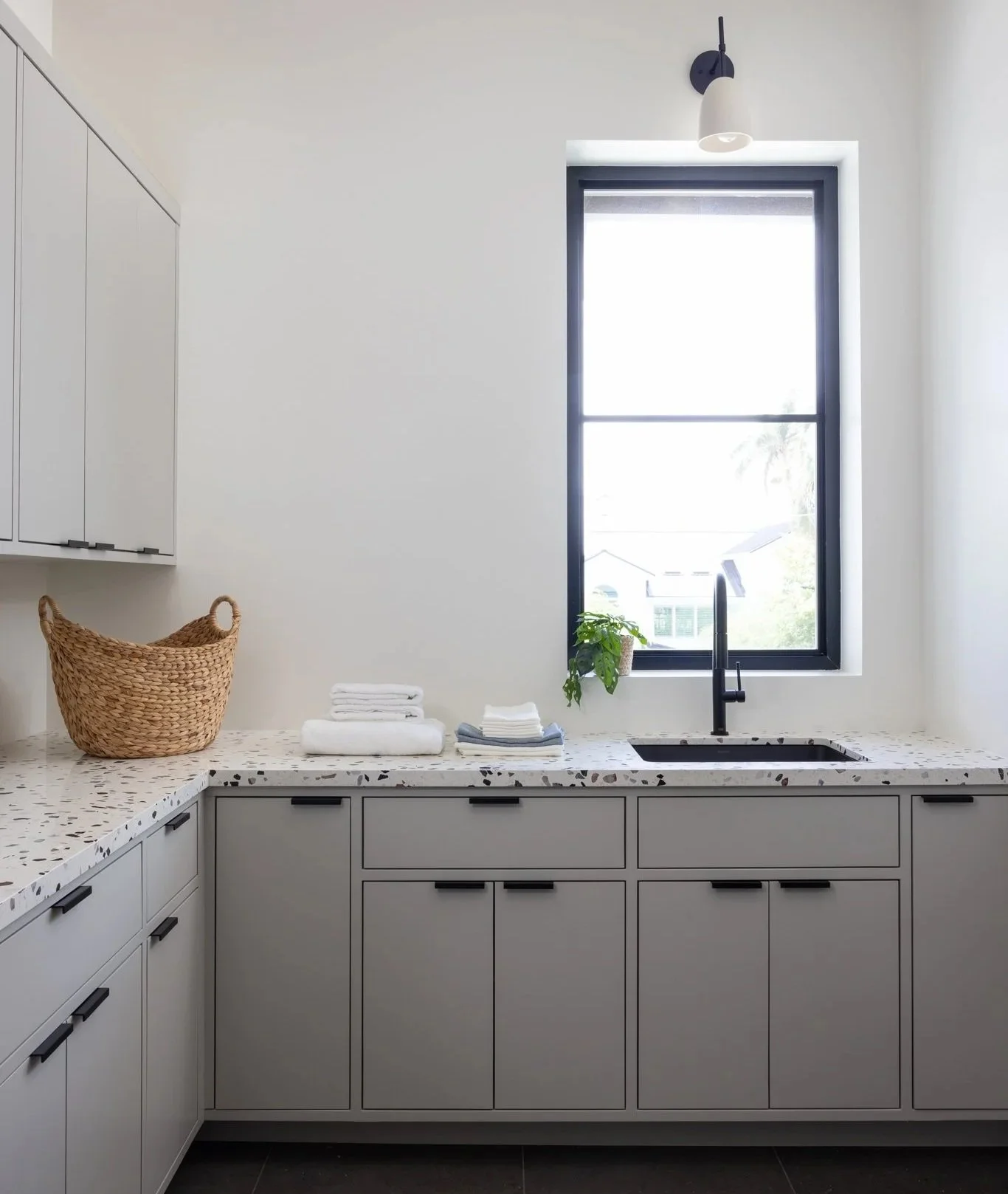 Modern laundry room with white cabinets, a speckled terrazzo quartz countertop, a black Blanco sink with a matte black faucet, a large window with black trim, a small green potted plant, a basket, and folded towels.