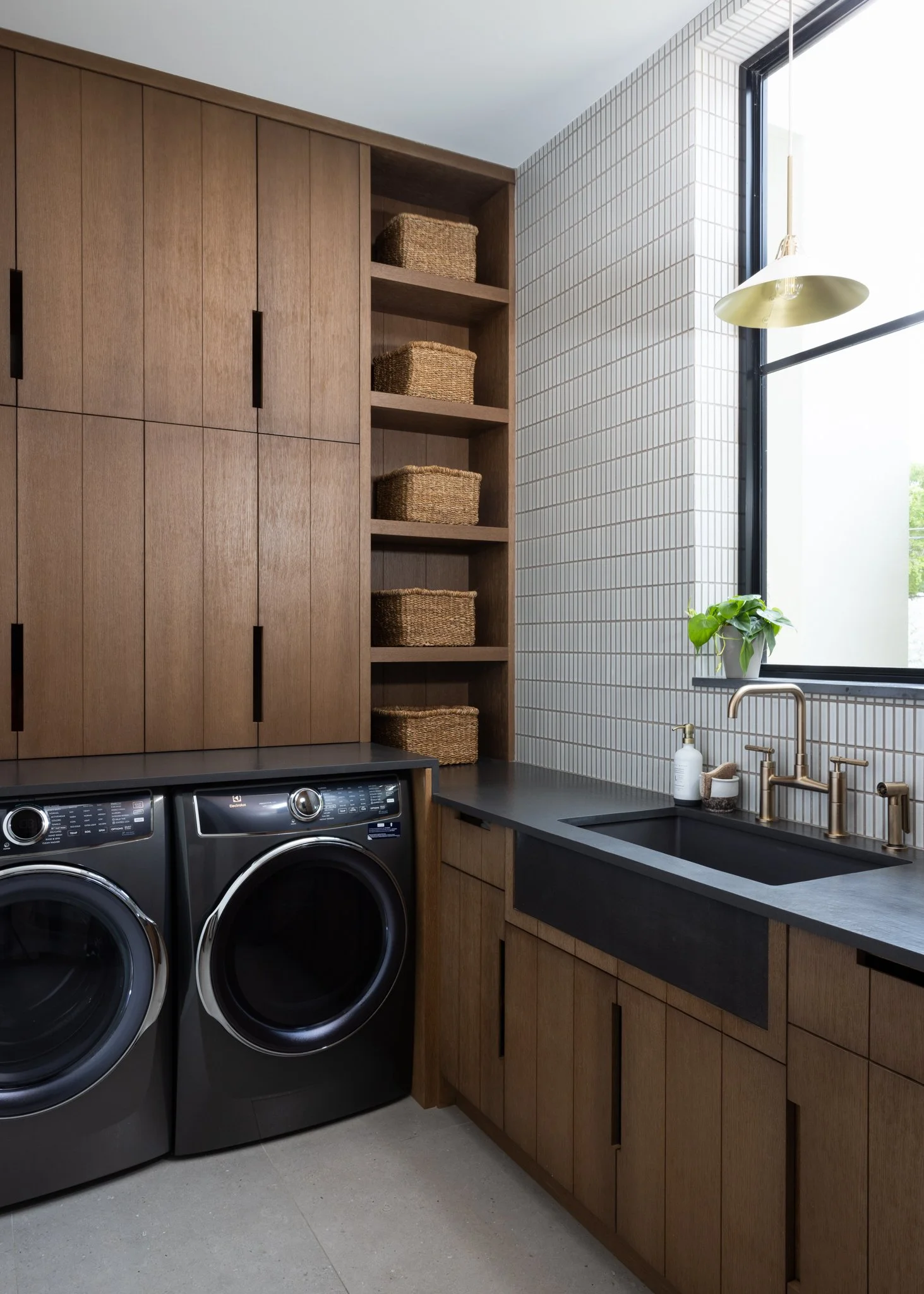 A modern laundry room with a black washer and dryer, wooden cabinetry, open shelves with wicker baskets, a black countertop, a black sink, a brass faucet, a white bottle, a small potted plant, a window with a black frame, and a gold pendant light.