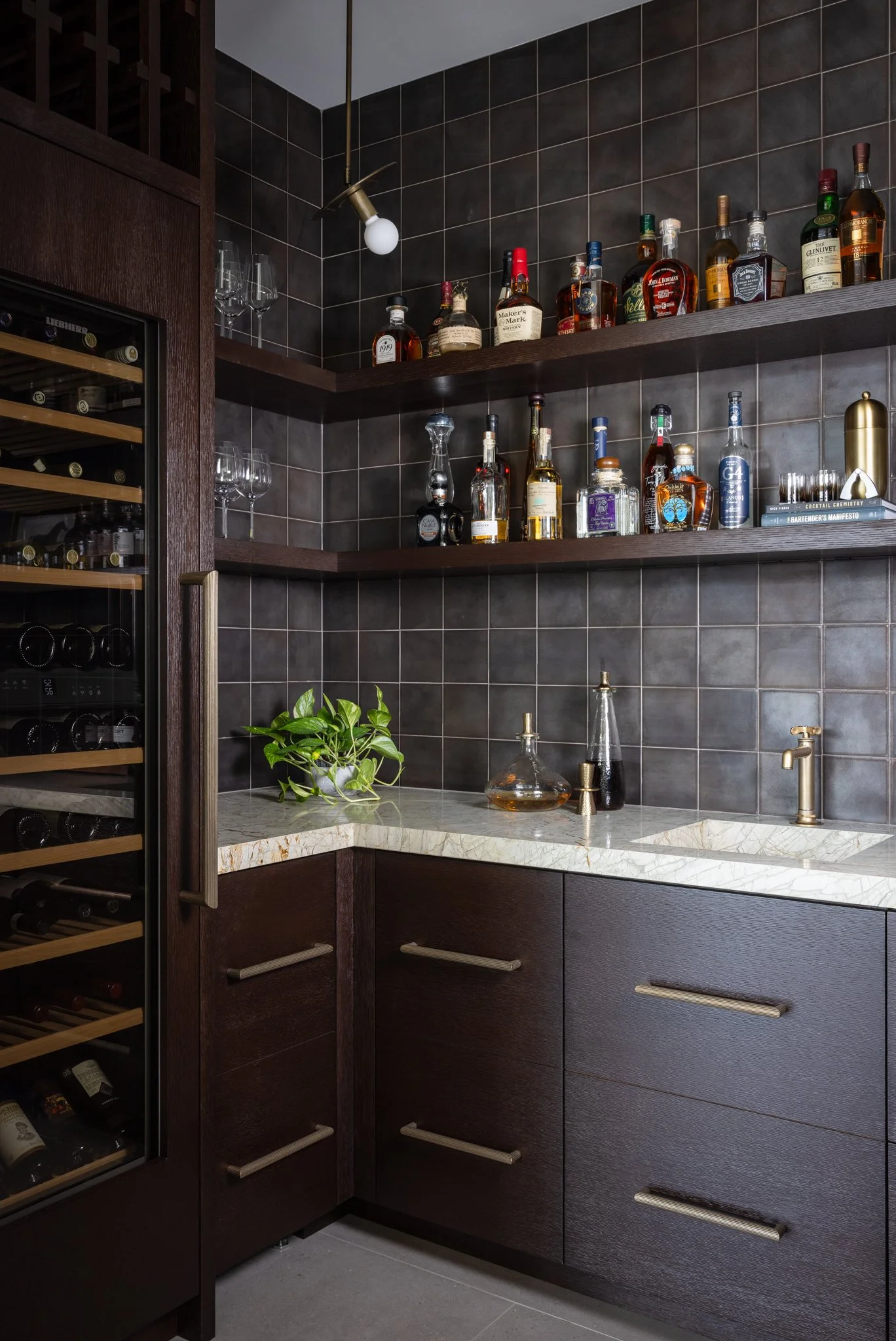 Modern bar area with dark wood cabinets and shelves, containing various bottles of liquor. A countertop with a marble surface features glass decanters and a plant. A wine fridge is visible to the left, and the background features dark tiled walls and