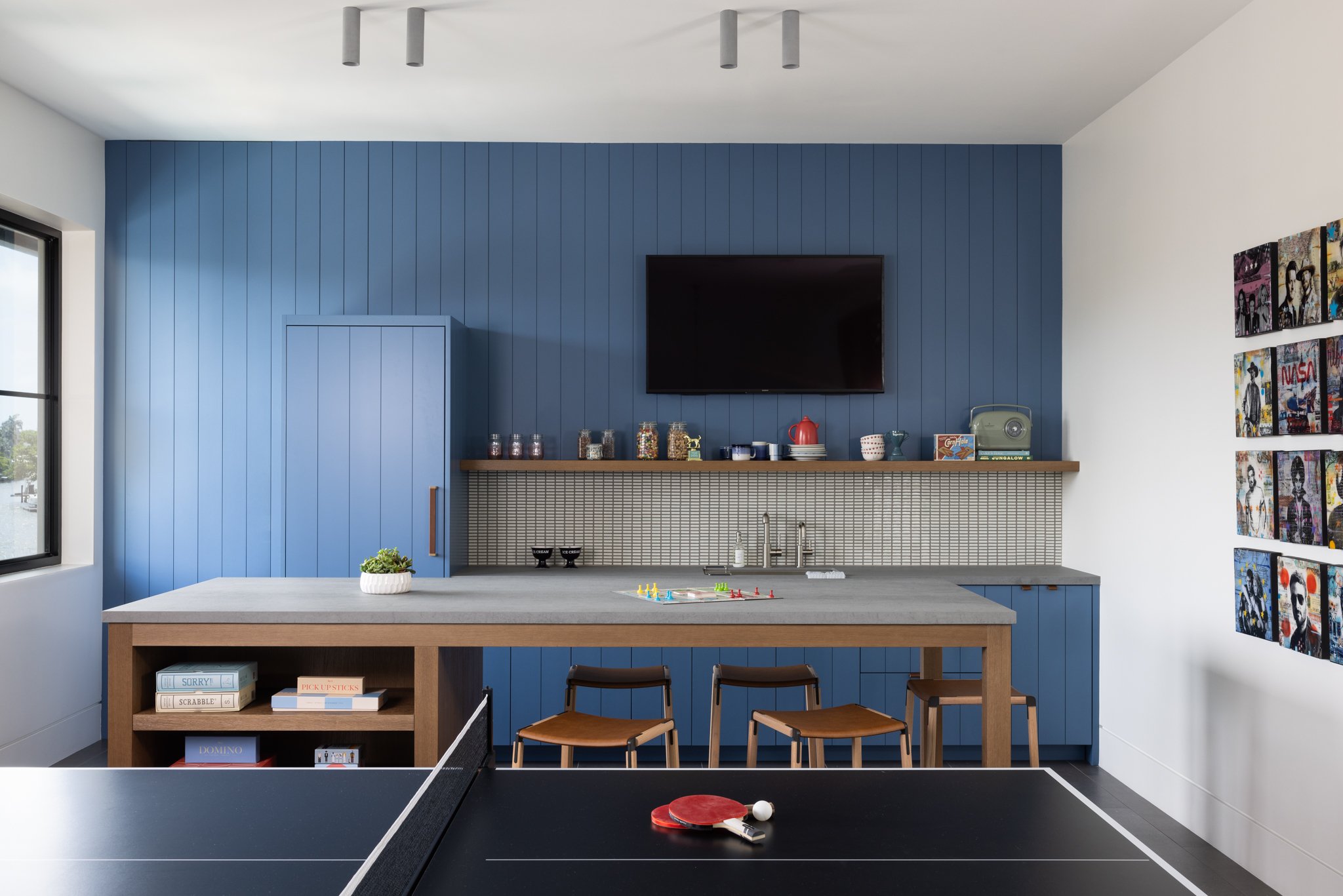 Modern teen play game room with blue paneled wall, TV mounted with integrated panel-ready appliances, a shelf with jars, cups, and books, and a table with ping pong paddles and ball, near a window overlooking rio vista neighborhood. 