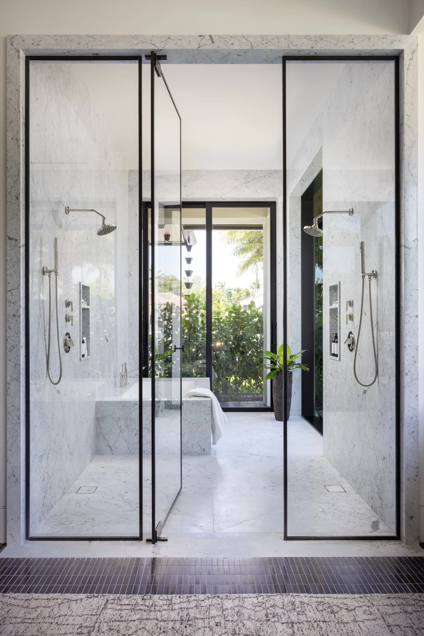 A modern wet room bathroom features a marble shower with two shower configurations from waterworks, a custom glass shower door, and a view of greenery outside through a large window.