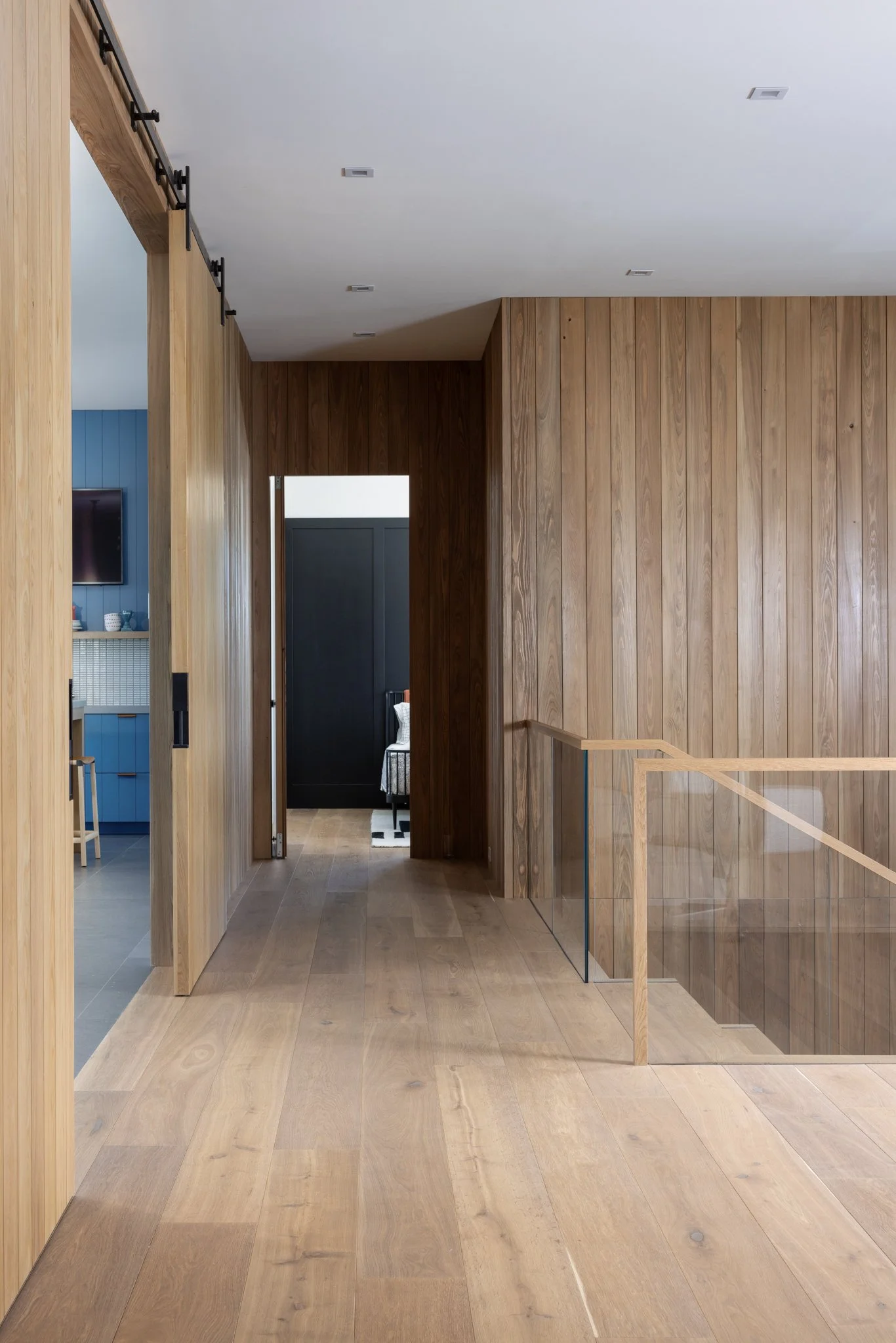 Interior of a modern house with natural wood walls and European wood flooring, featuring a hallway with glass railing with a view into a game room and guest room with black walls and striped rug. 