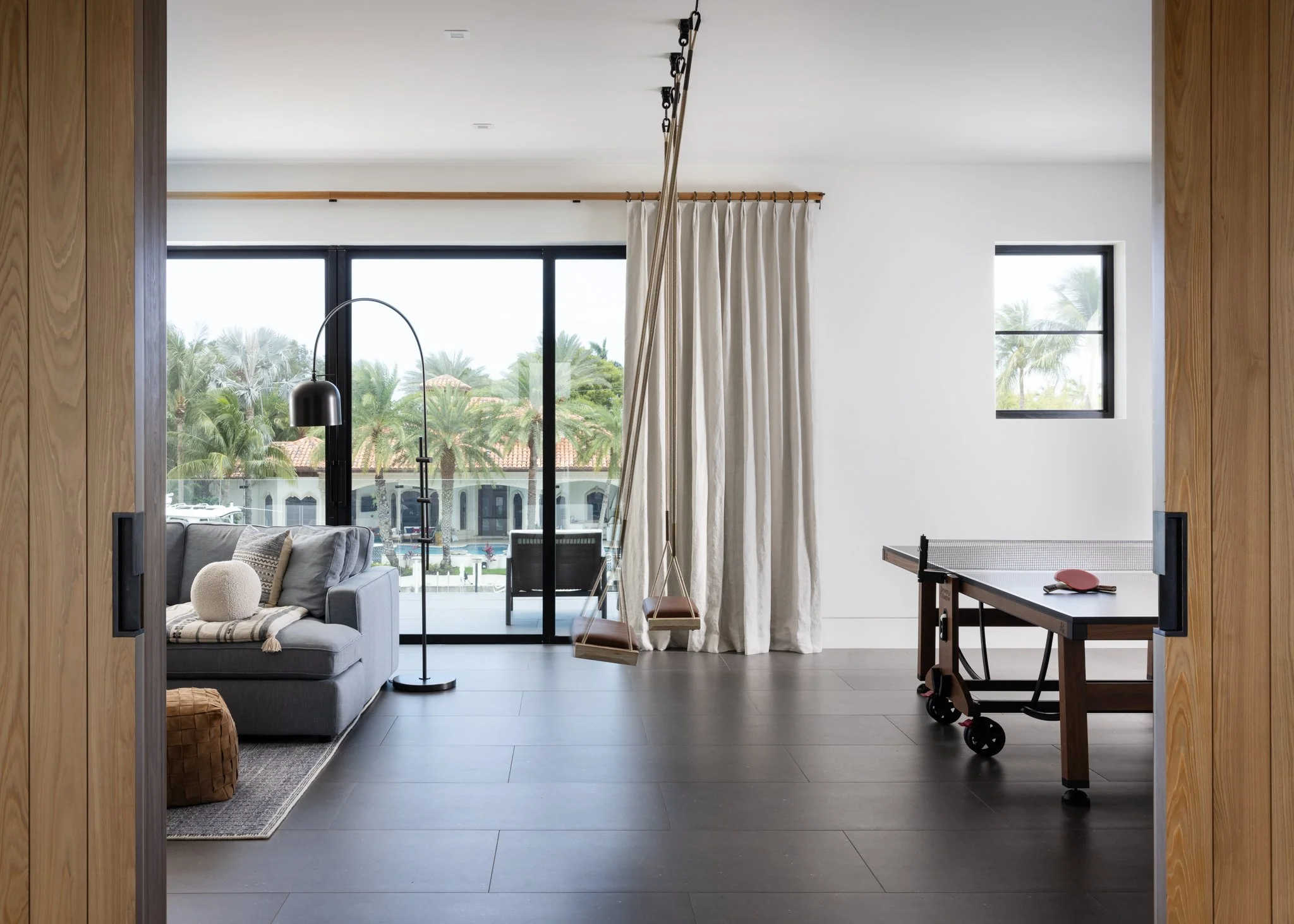 Modern game room with Rocce Fumo tiles from Opus Stone large sliders showing palm trees in Fort Lauderdale with a custom interior define sofa, floor lamp, custom curtains and rod, a ping pong table, and interior swings by Buzzell Studios. 