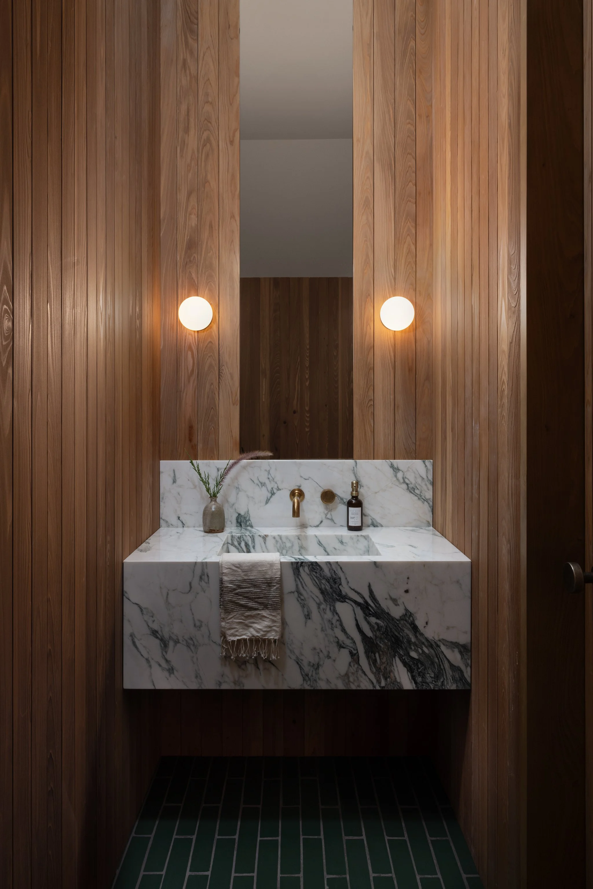 A modern bathroom with wooden panel walls under the stairs, a floating white corchia marble sink with black veins, gold allied maker orb ixtures, two round wall lights, a small vase with green plants, and dark wow tiles flooring.